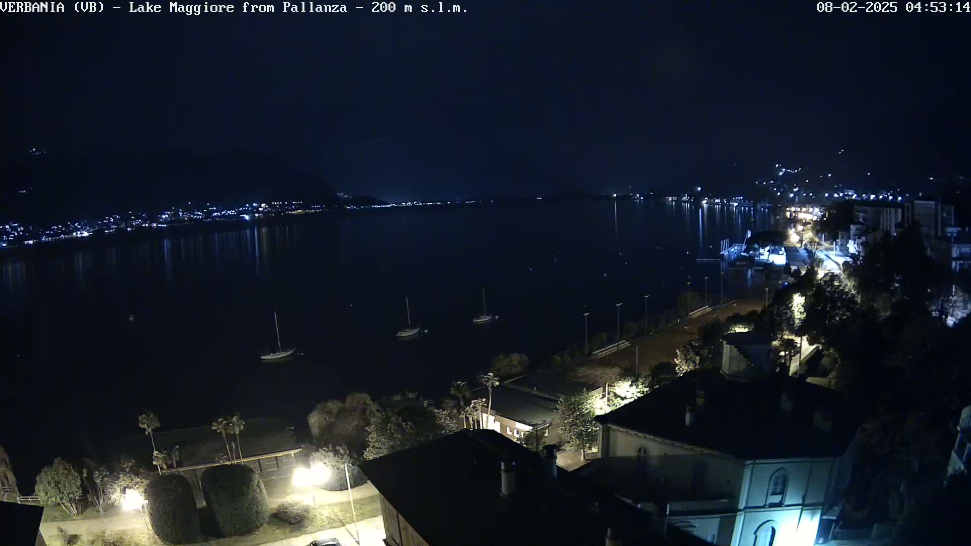 A nighttime view of a calm lake reflecting city lights, with a few sailboats moored near a shoreline lined with buildings and trees.