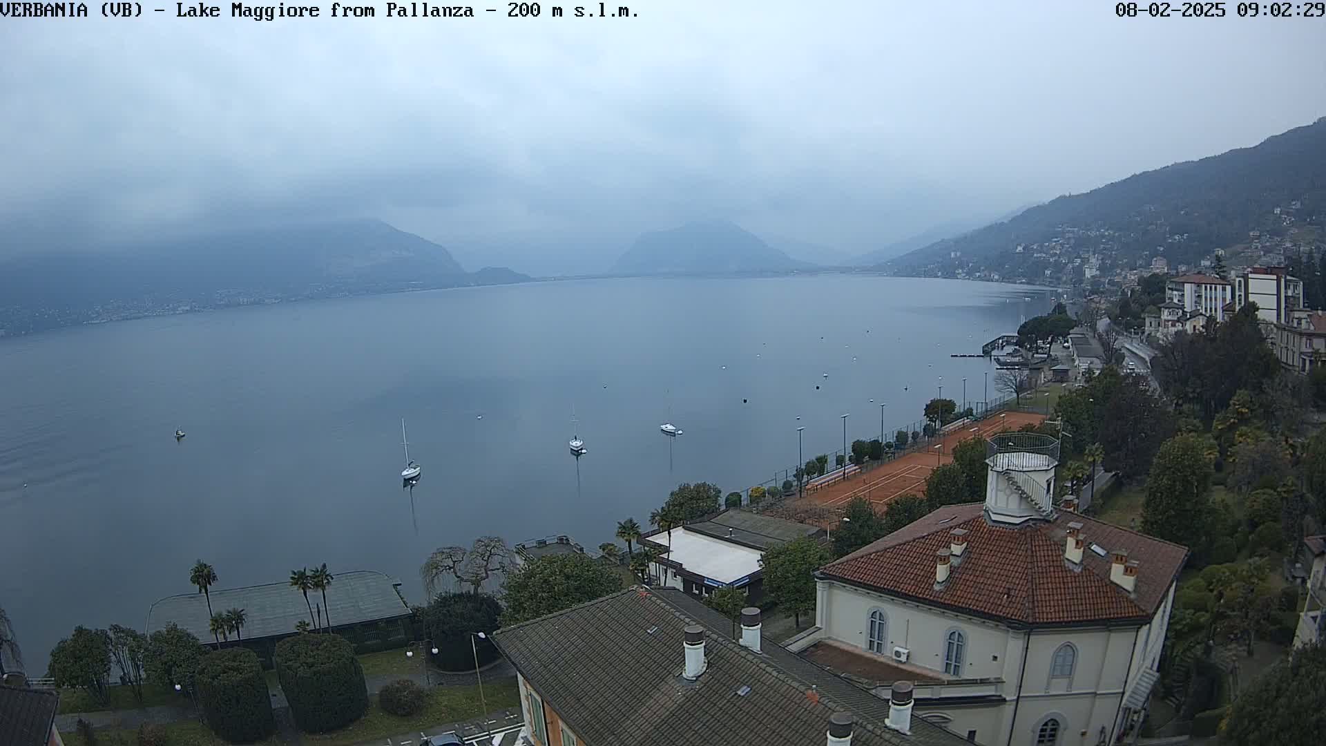 A hazy, overcast day reveals a view of Lake Maggiore, with several small boats on the water, a tennis court, and buildings situated along the shoreline.