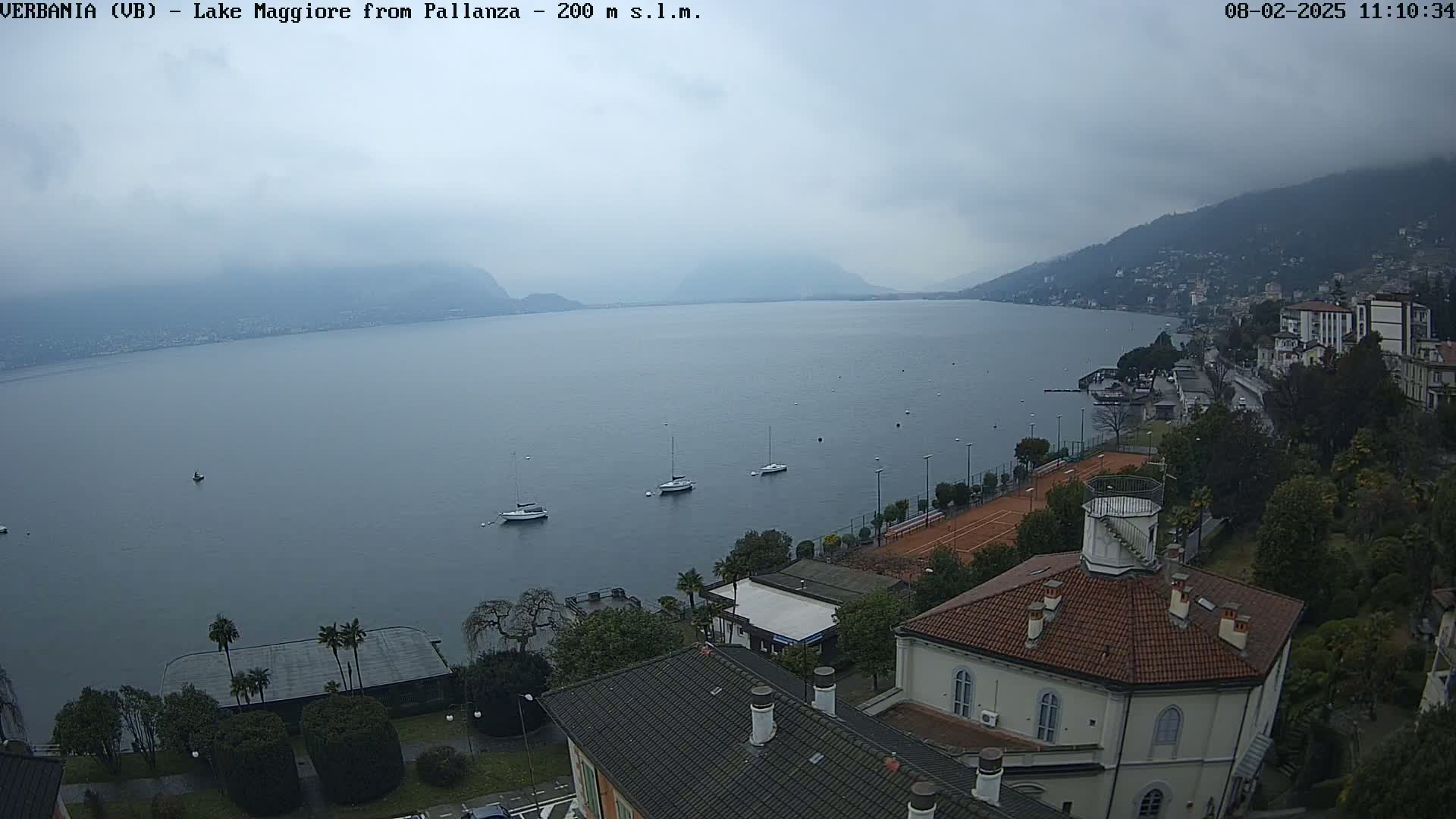 A cloudy day overlooks a calm lake with several sailboats and a lakeside town, featuring buildings with red-tiled roofs and tennis courts.