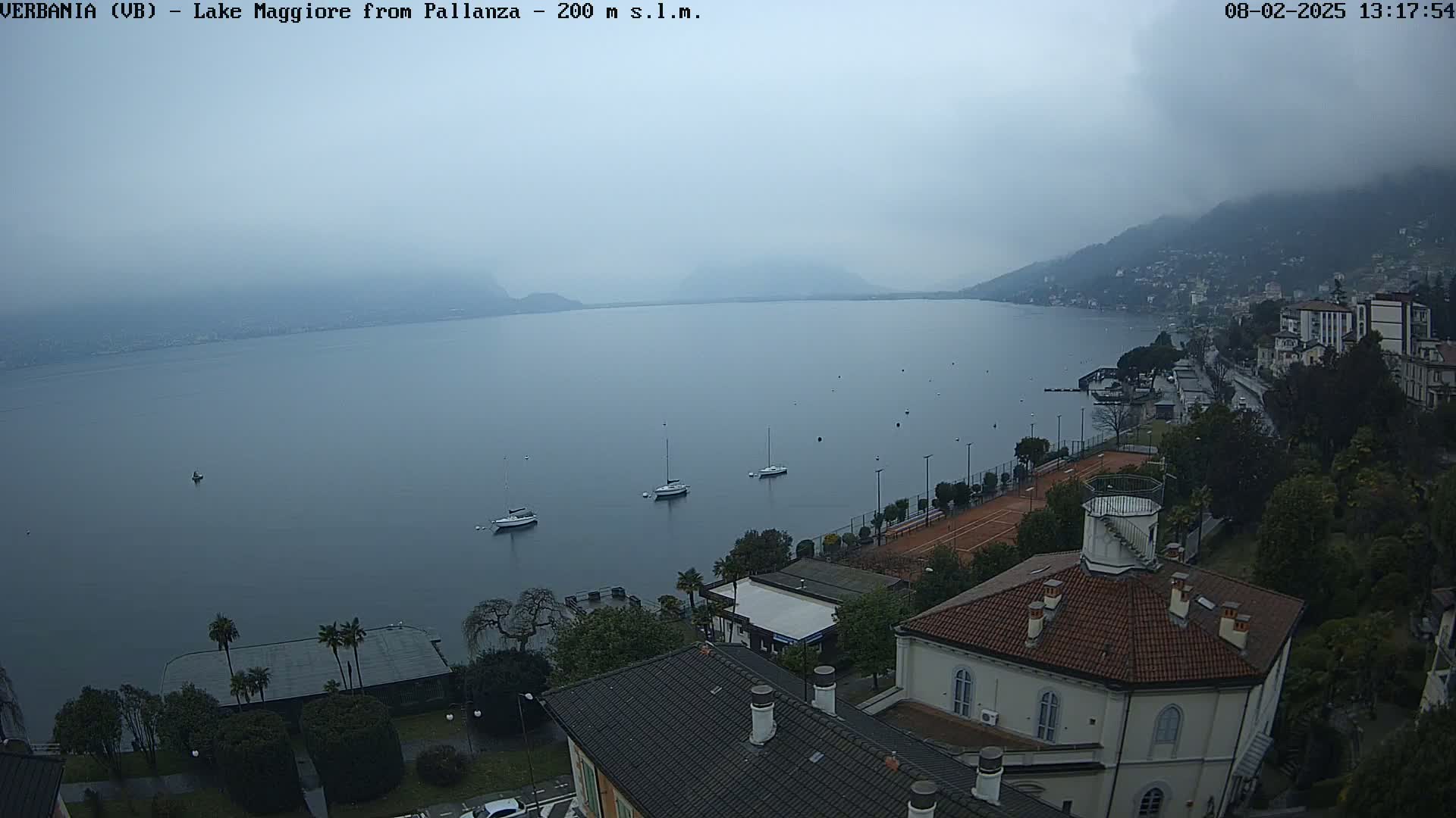 A hazy, overcast day reveals a calm lake with several sailboats, a shoreline with buildings and trees, and a tennis court; in the foreground is a building with a red roof.