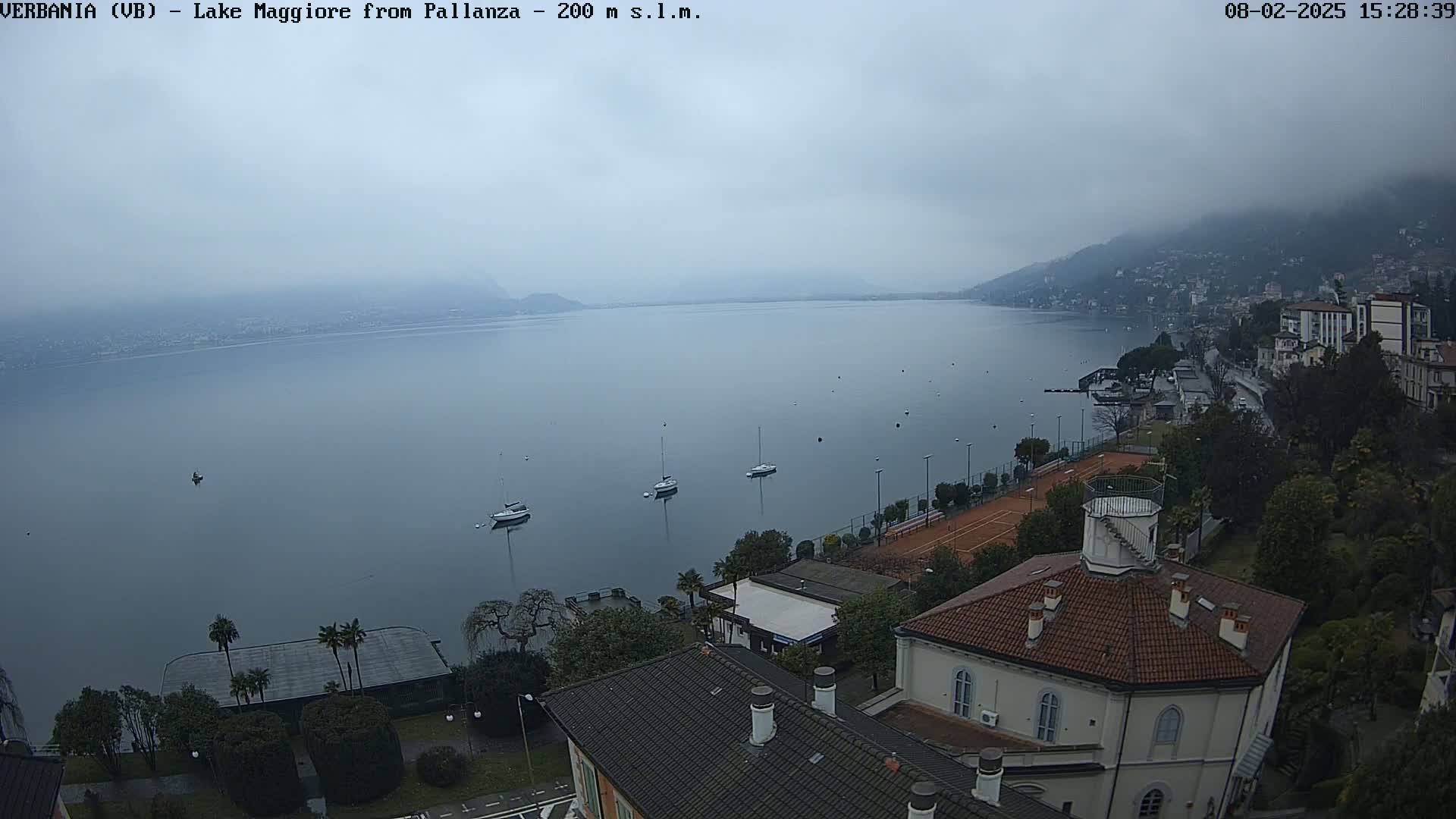 A foggy view of a lake with several sailboats, tennis courts, and buildings on a shoreline.