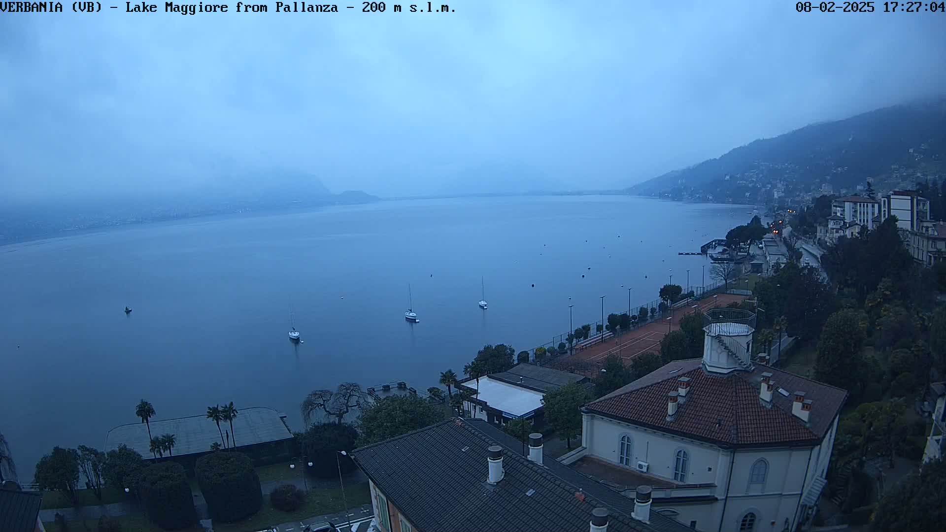 A hazy, overcast view of a lake with several sailboats, a shoreline town, and buildings, including a building with a round roof and a tennis court.