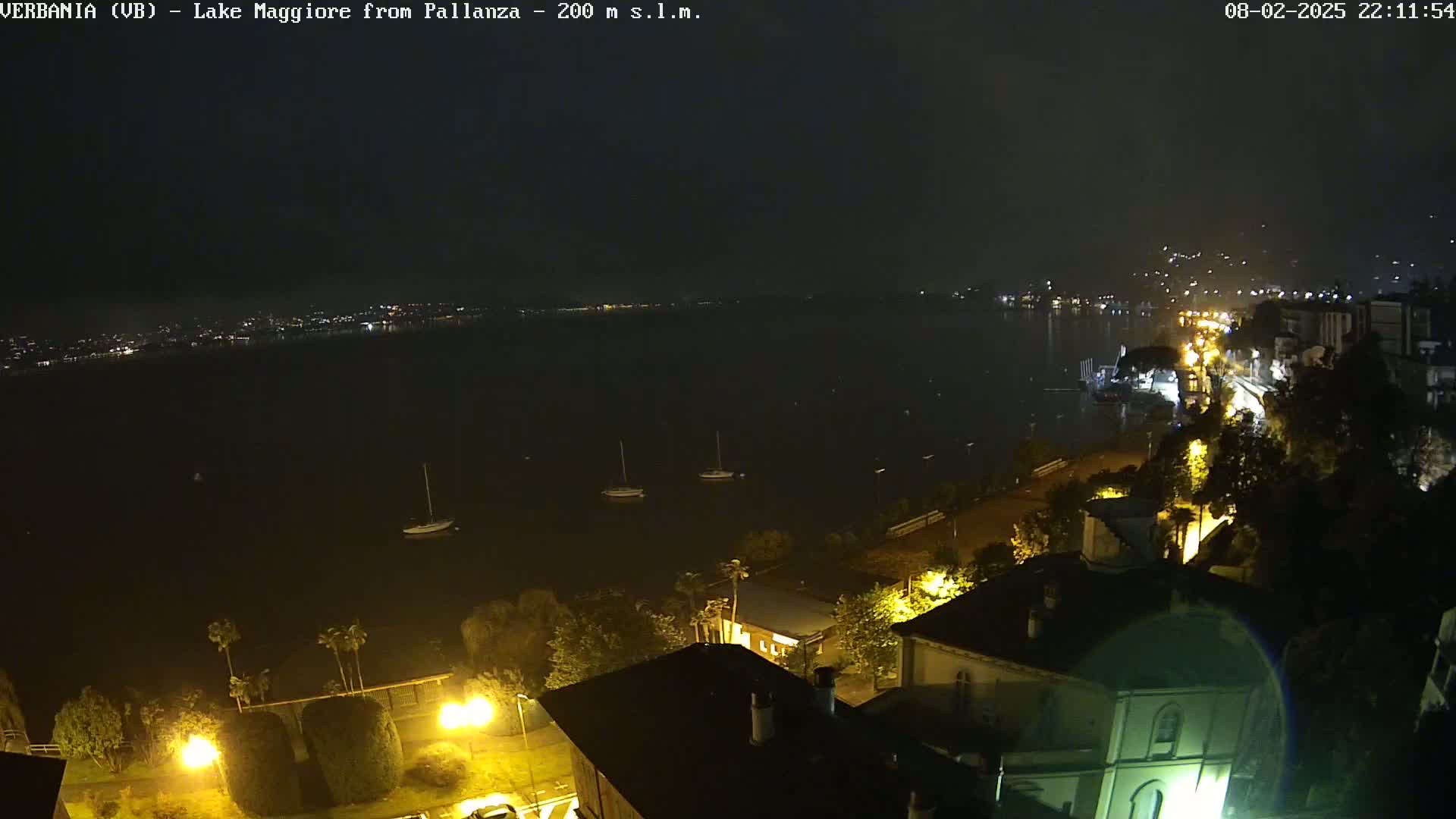 A nighttime view of a lake with a few boats, a shoreline with buildings and streetlights, and a city across the water.