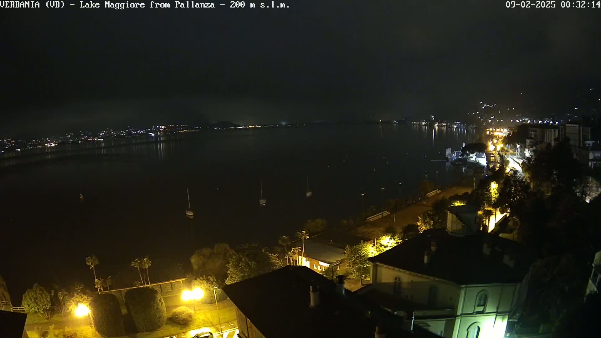 A nighttime view of a lake with a town's lights visible along the shore under a dark, cloudy sky.