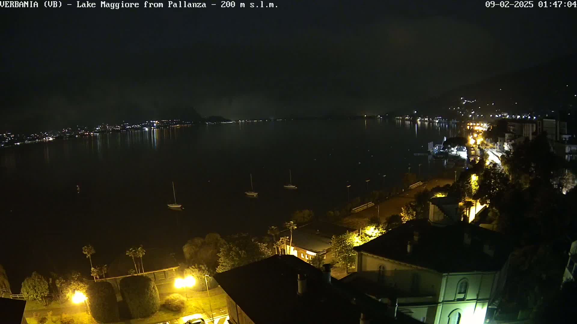 A nighttime view of a calm lake with a few boats, a town with lights illuminating buildings and streets along the shoreline, and hills in the background under a cloudy sky.
