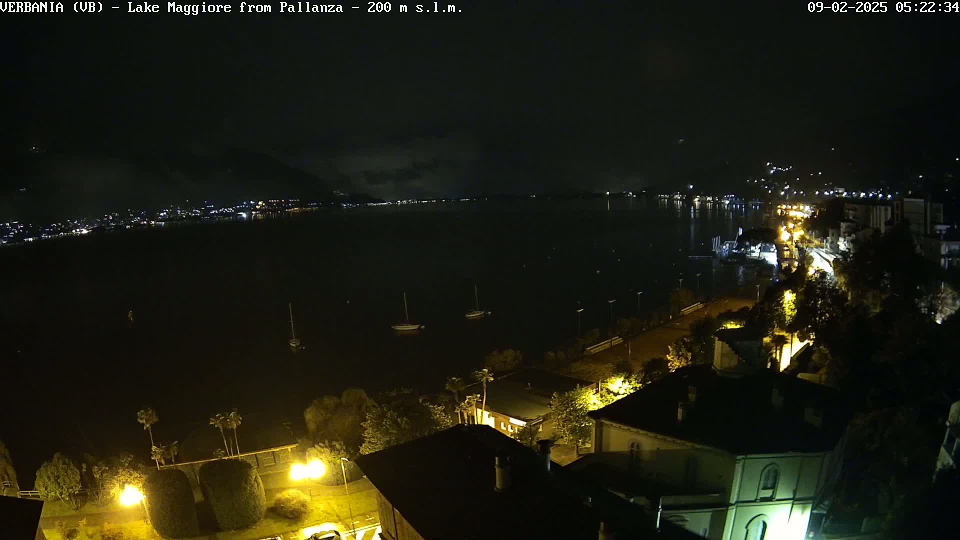 A nighttime view of a lake, with a town's lights visible along the shore and a few sailboats on the water.