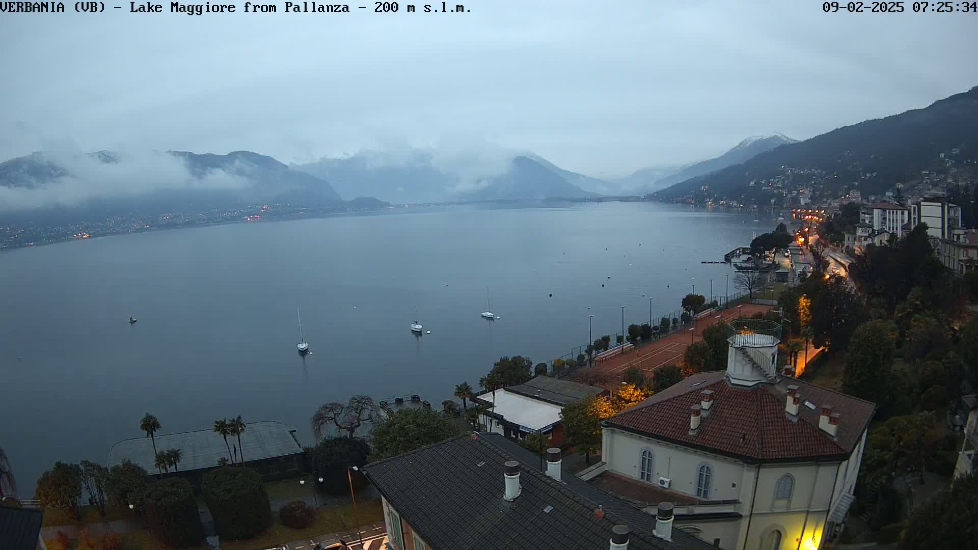 A calm lake is viewed from a hilltop overlooking a town at dawn under an overcast sky, with a few sailboats visible on the water.