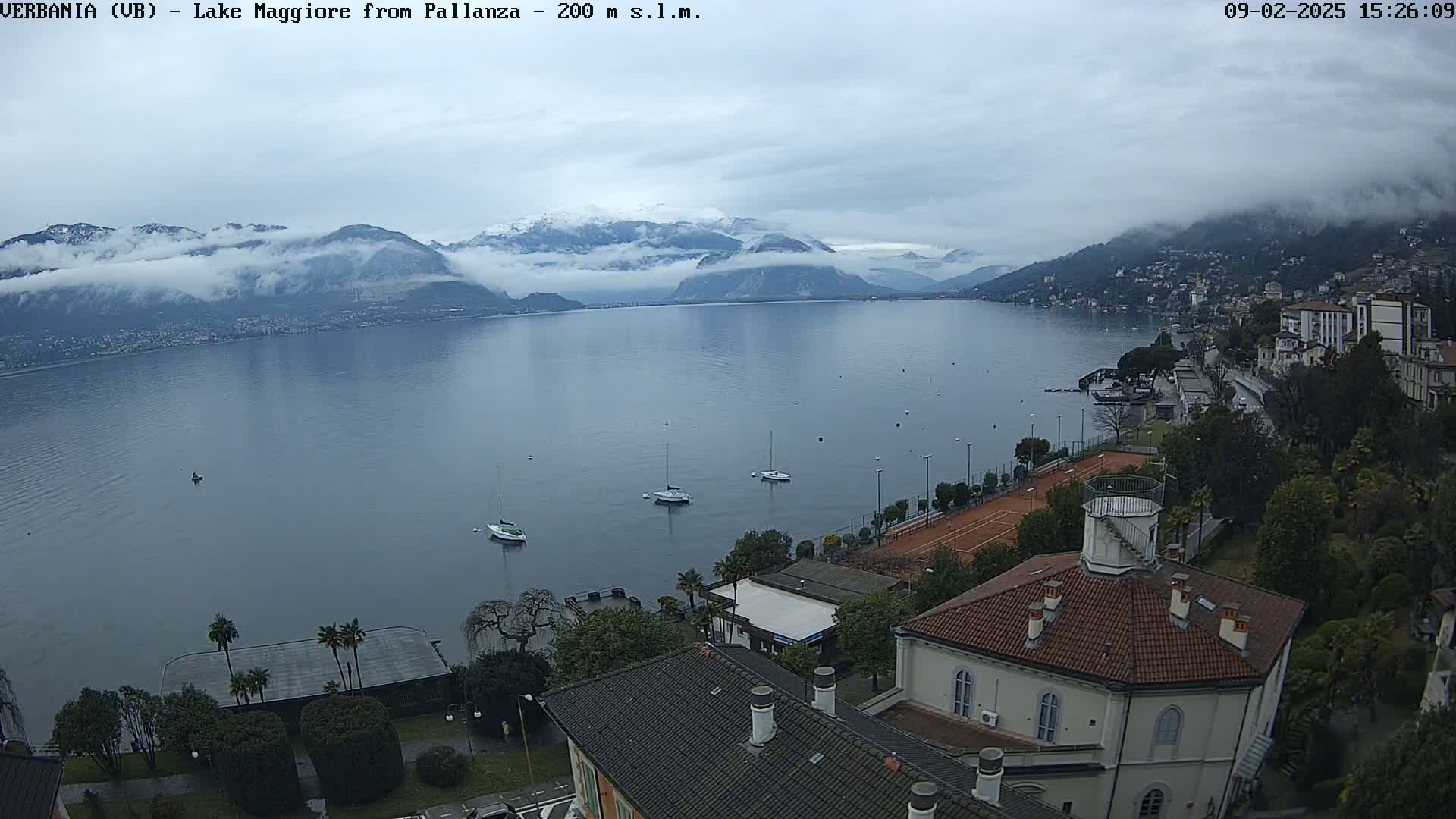 A high-angle view shows a calm lake with several sailboats, surrounded by buildings, trees, and mountains partially obscured by fog under an overcast sky.