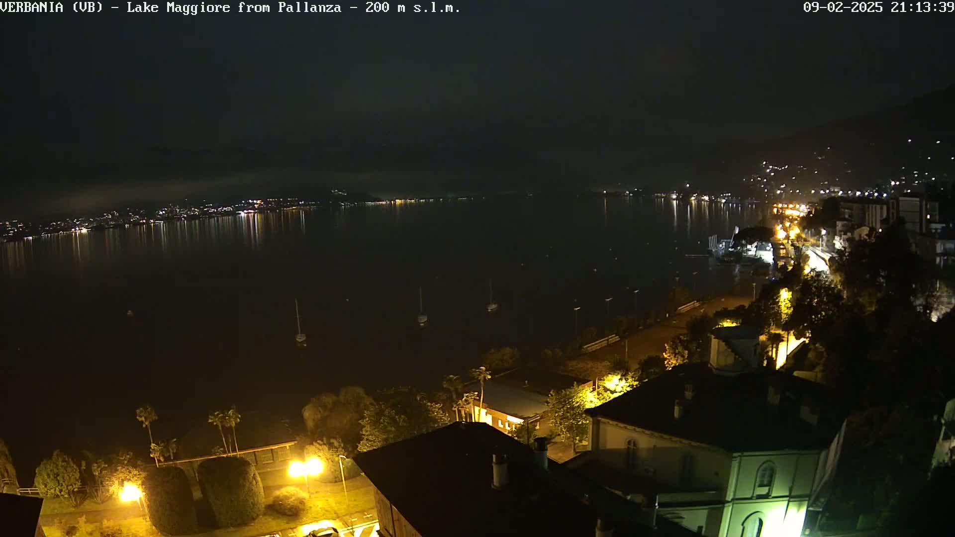 A nighttime view of a lake reflecting city lights, with several boats visible on the water and illuminated buildings and trees along the shoreline.
