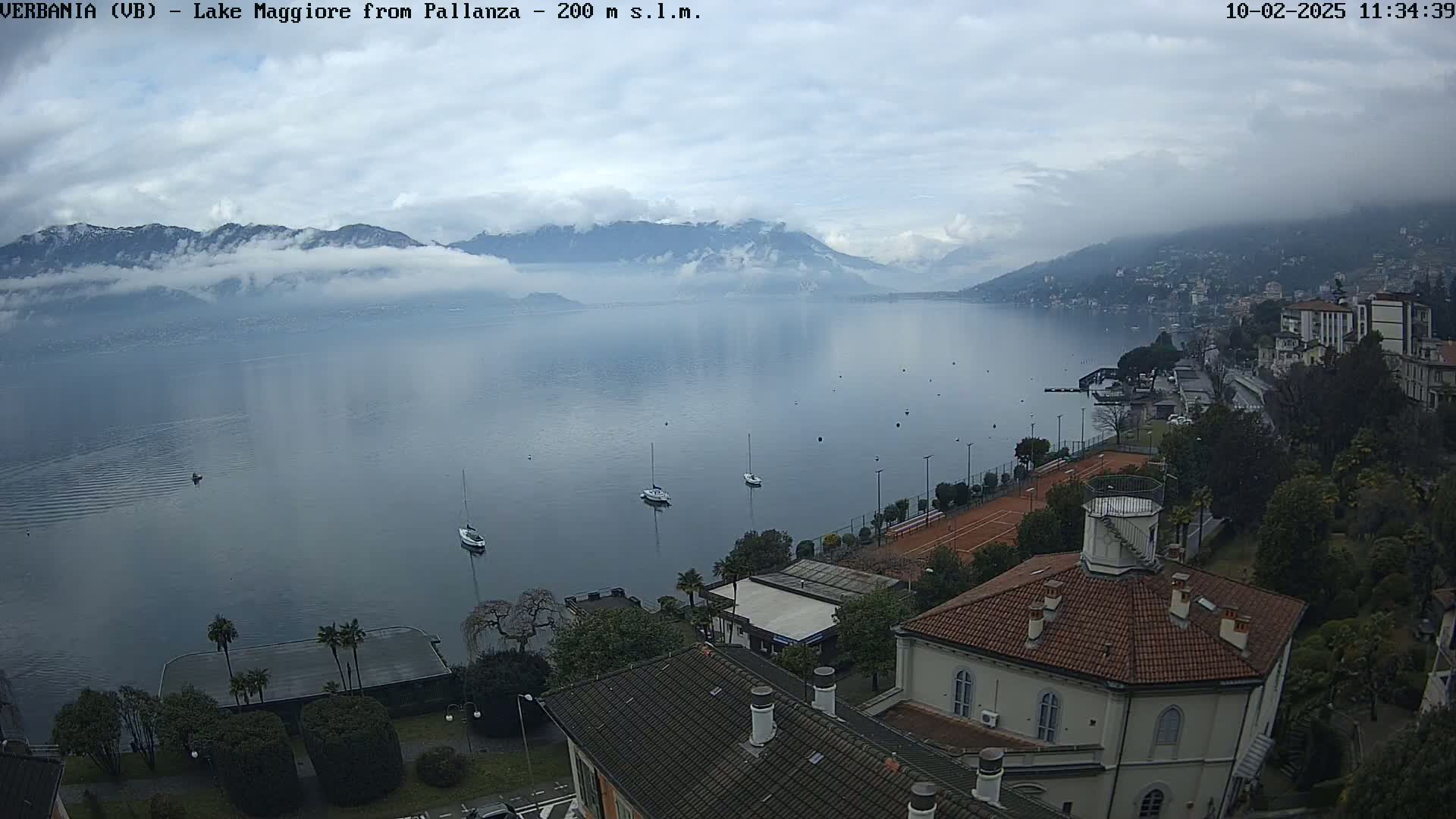 A calm lake with several sailboats is surrounded by buildings, trees, and mountains partially obscured by fog under an overcast sky.