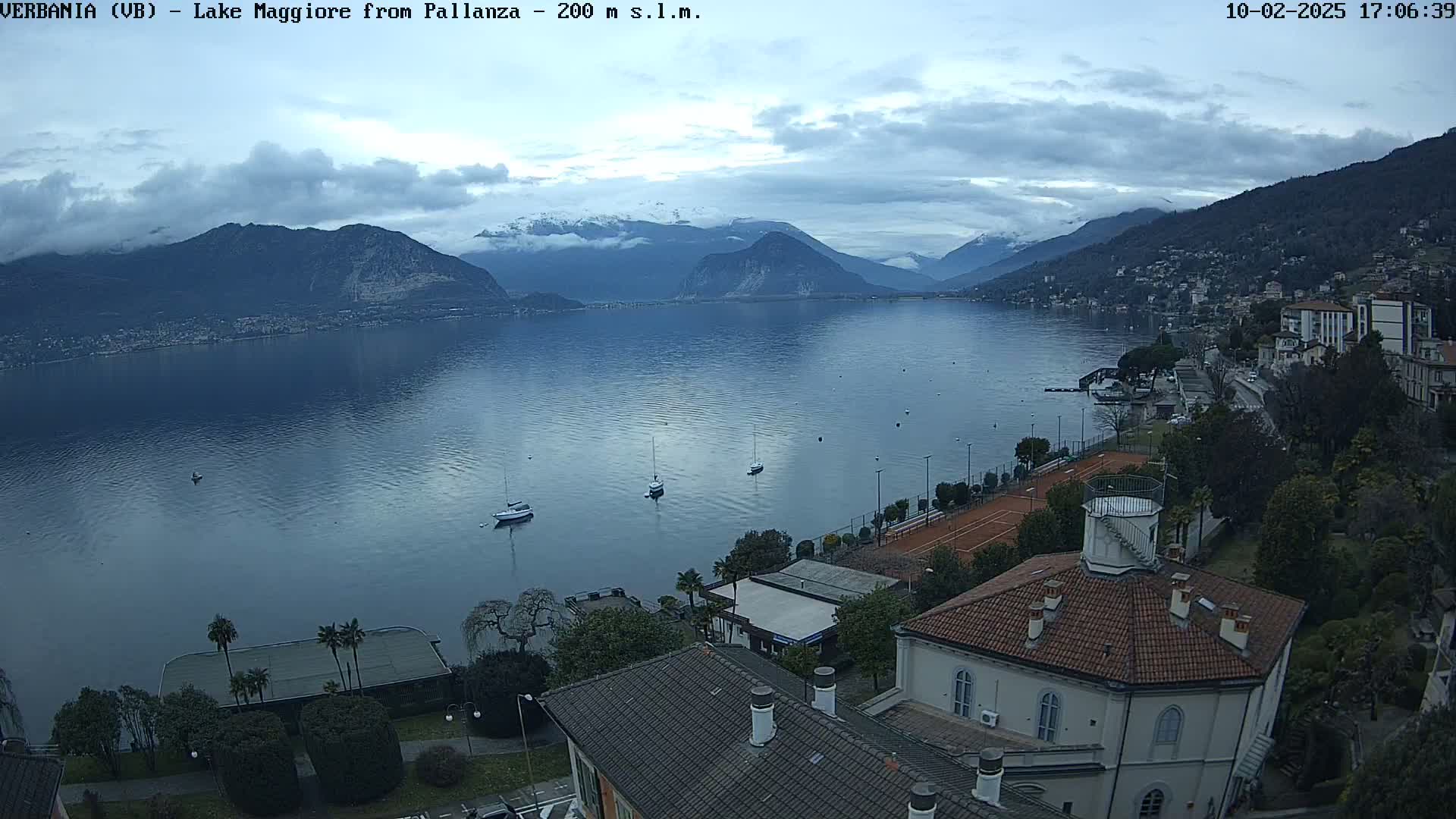 A calm lake with several sailboats is nestled between snow-capped mountains under a cloudy sky, overlooked by buildings and tennis courts.