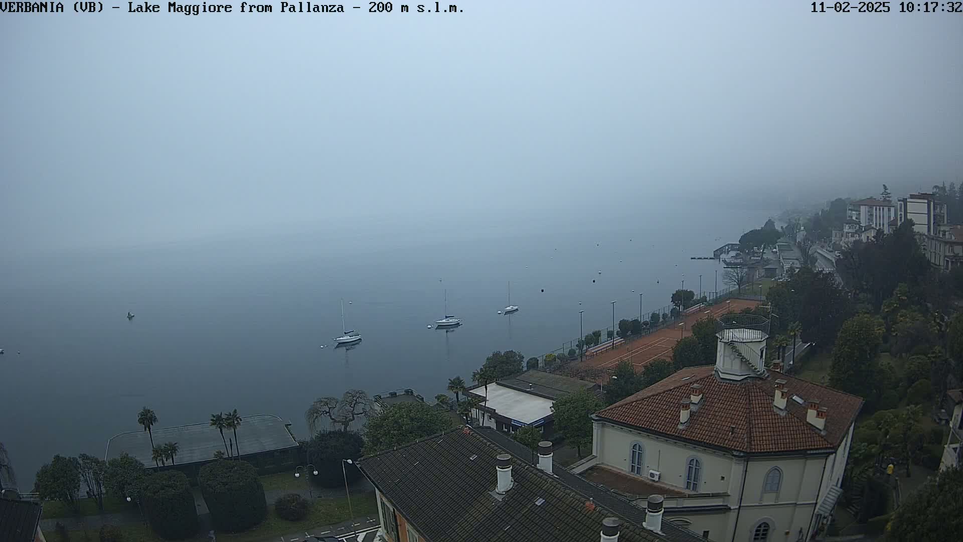 A foggy view of a lake with several boats and a shoreline featuring buildings, tennis courts, and lush greenery.