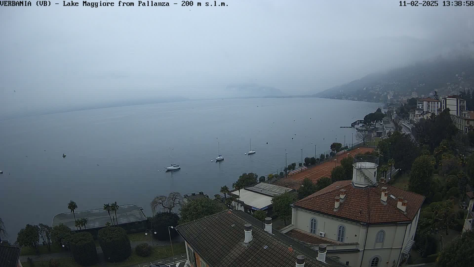 A hazy, overcast day reveals a calm lake with several sailboats, a shoreline with buildings and a tennis court, and a view from a slightly elevated vantage point.