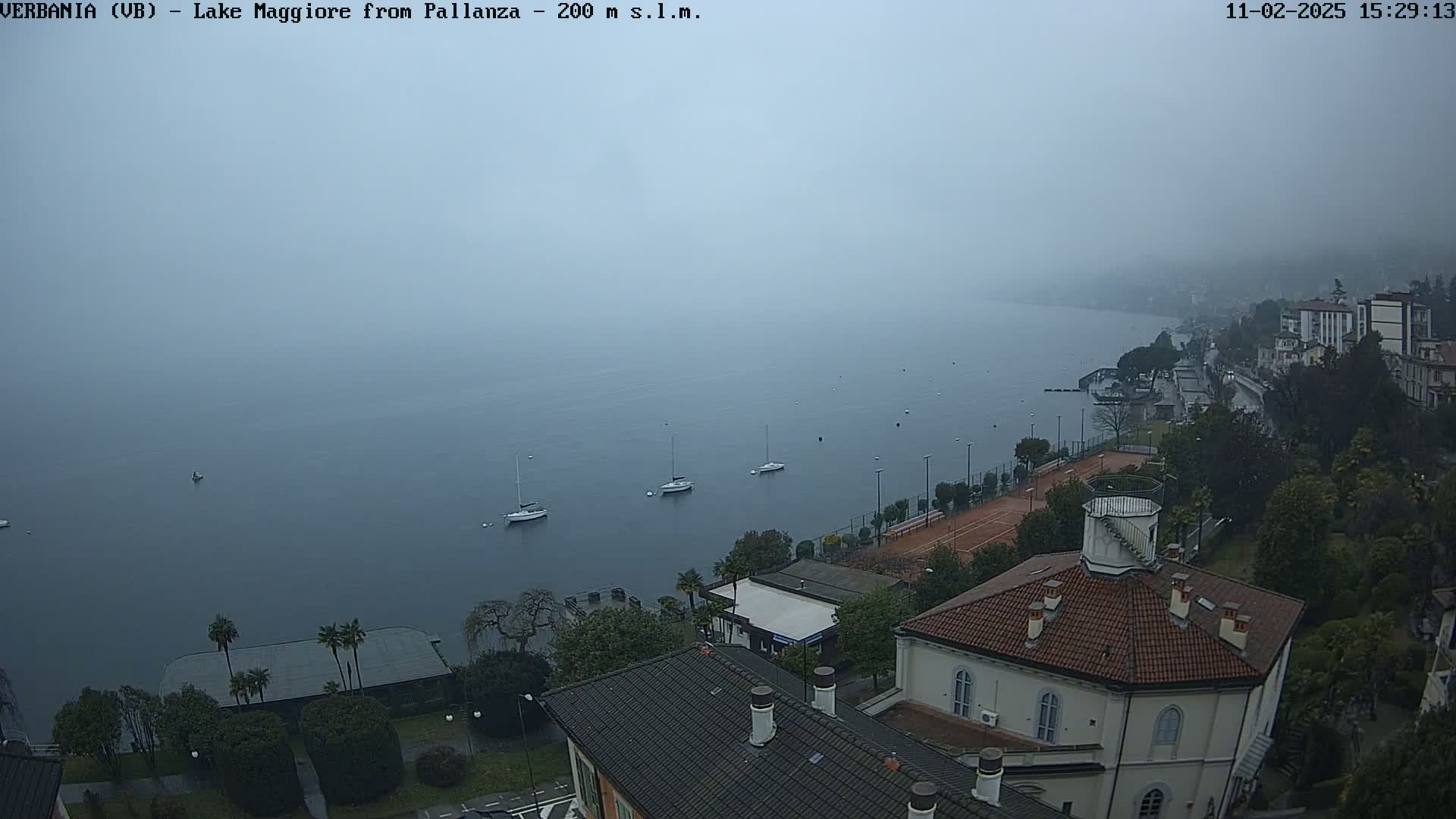 A foggy view of Lake Maggiore from a high vantage point, showing several sailboats on the water and a lakeside town with buildings and greenery.