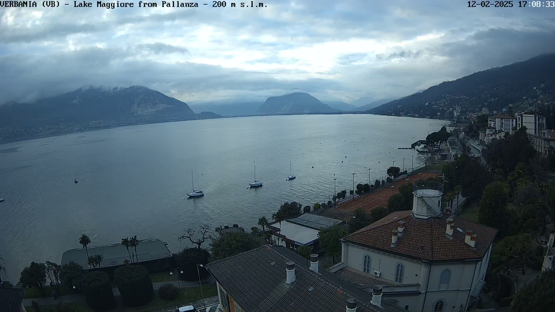 An overcast day reveals a tranquil lake scene with several sailboats, tennis courts, and buildings nestled against a mountain backdrop.