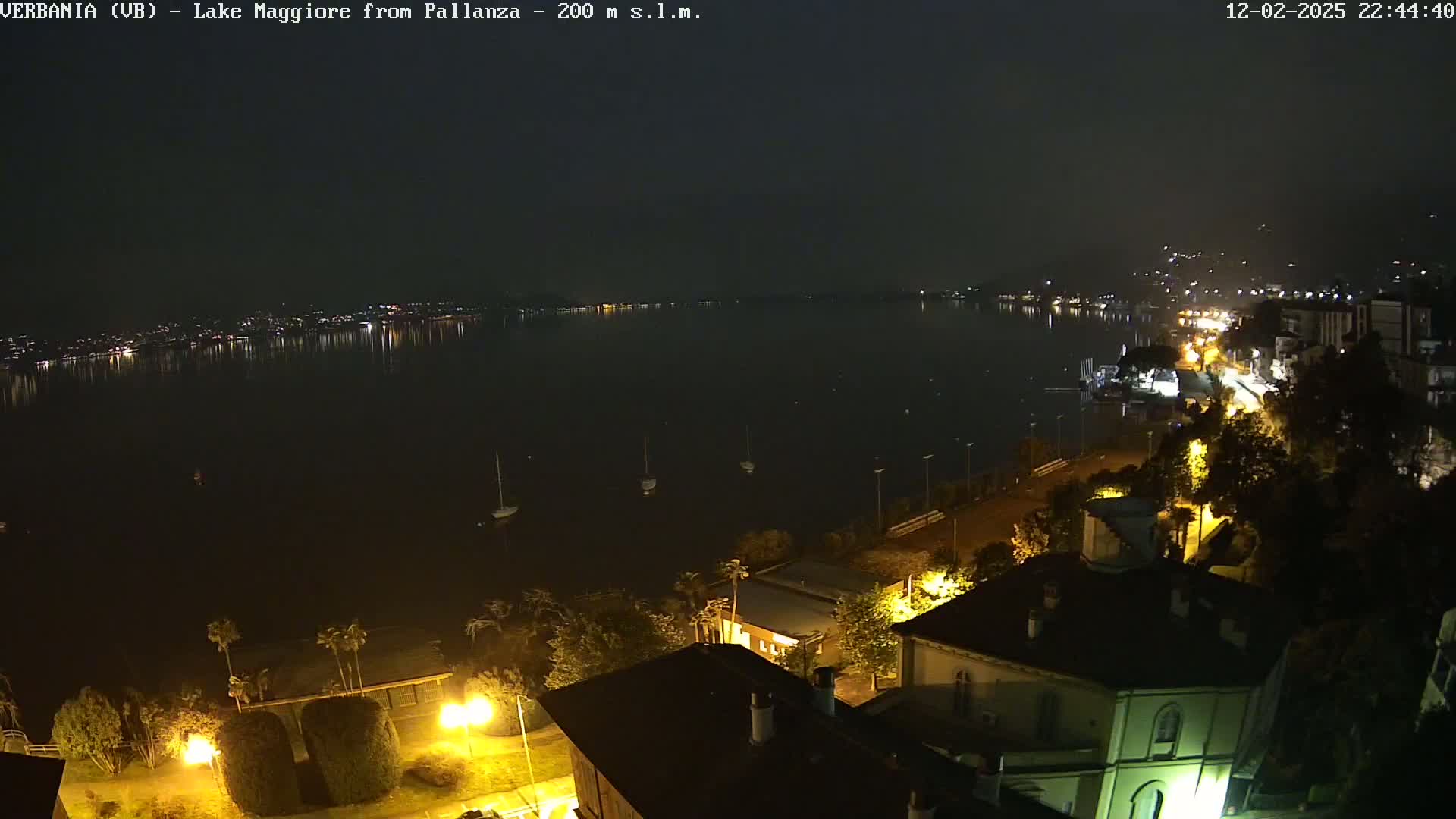 A nighttime view of a calm lake with lights reflecting on the water, a shoreline with buildings and streetlights, and a few boats.