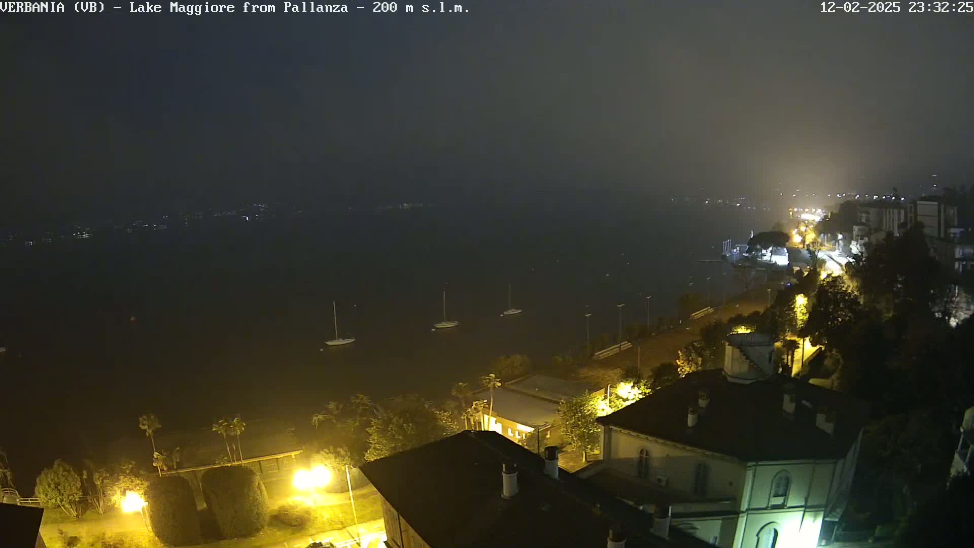 A nighttime, hazy view of a lake with several sailboats, and a lakeside town with illuminated buildings and streetlights.