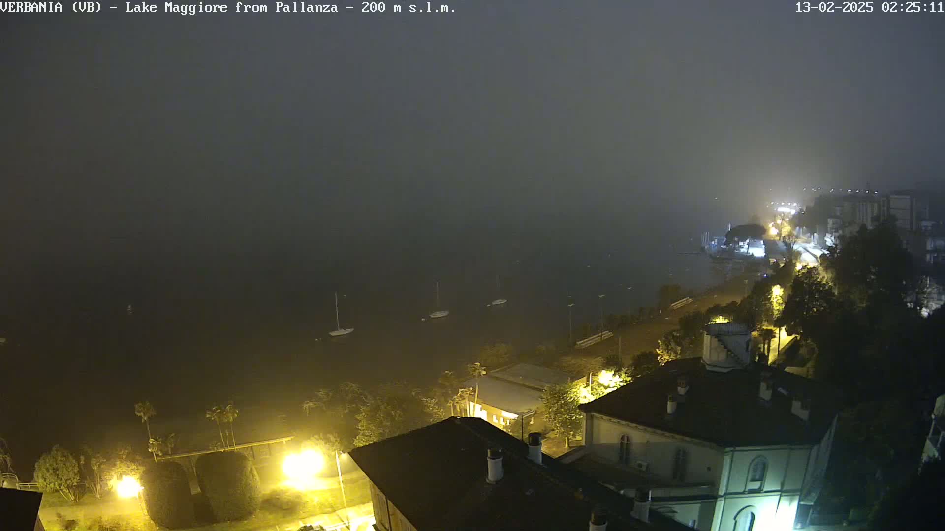 A foggy night scene shows a lake with several sailboats, a shoreline with buildings and streetlights, all under a thick fog.