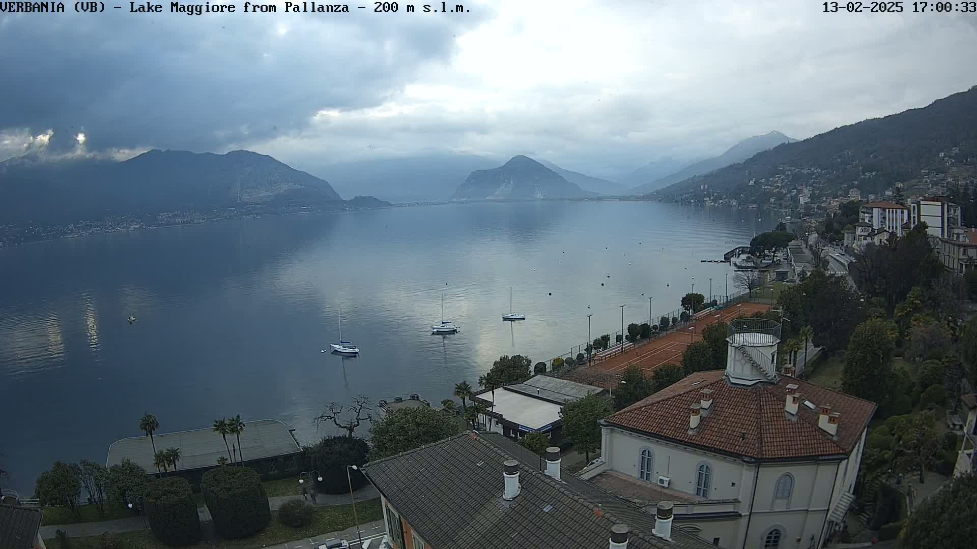 An overcast day reveals a tranquil lake surrounded by mountains and a lakeside town, with several sailboats visible on the water and tennis courts nestled among buildings.