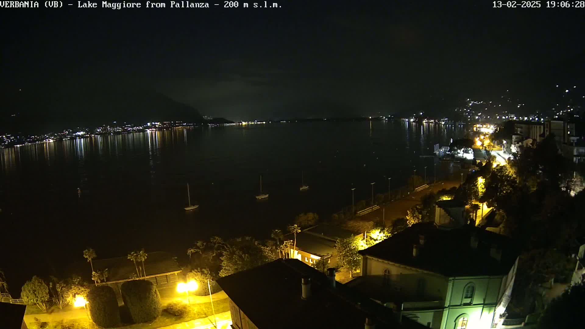 A nighttime view of a calm lake with lights reflecting on its surface from a town on the opposite shore, and a few boats moored near the foreground, under a clear dark sky.
