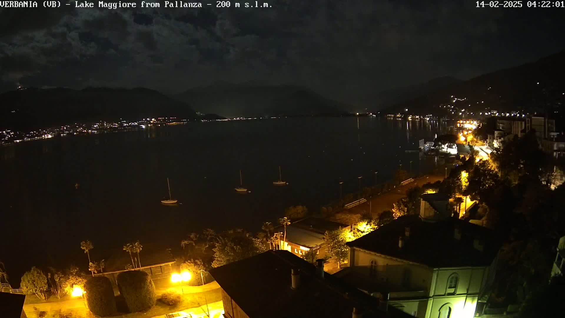 A nighttime view of a lake with a few boats, a shoreline with illuminated buildings and trees under a partly cloudy sky.