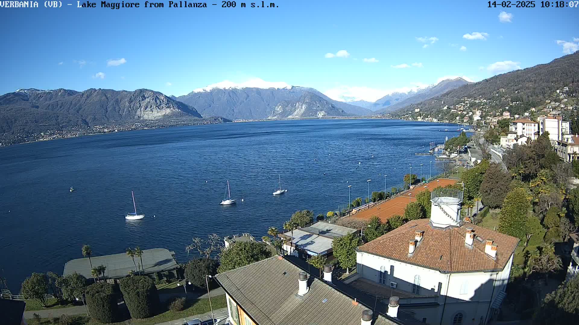 A sunny day shows a view of a lake with snow-capped mountains in the background, boats on the water, and buildings and tennis courts along the shoreline.