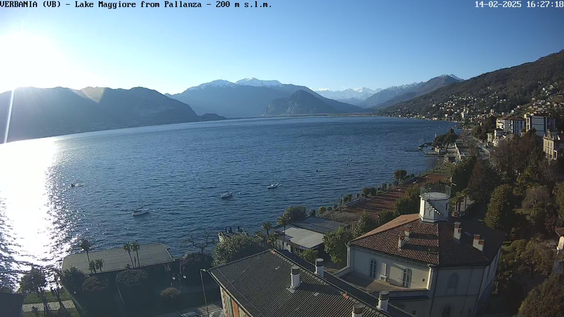 A sunny day overlooks a calm lake with snow-capped mountains in the background and a lakeside town with buildings and trees in the foreground.