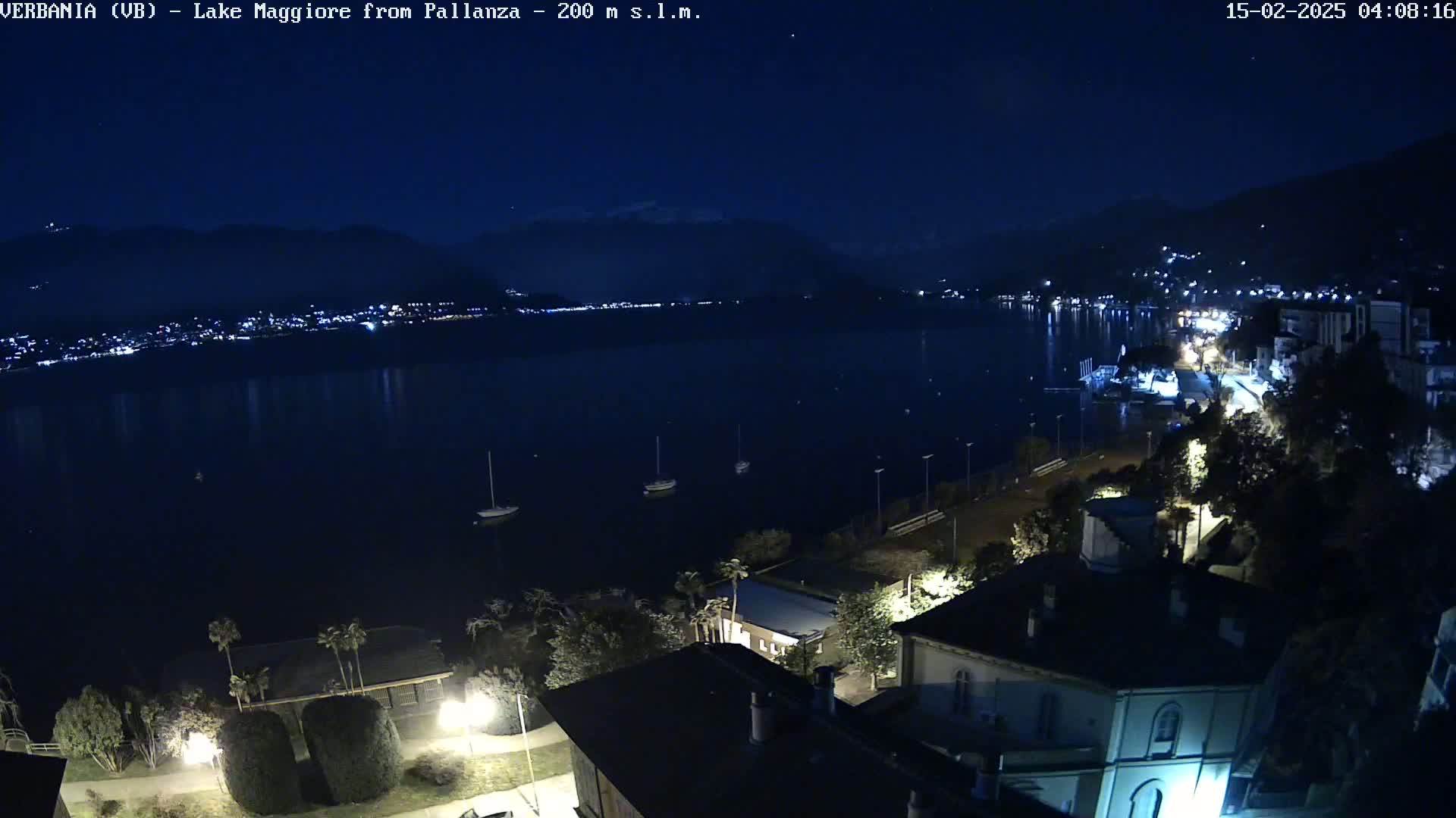 A nighttime view of Lake Maggiore from a hilltop overlooking a town, with several sailboats on the dark water and lights illuminating the shoreline.
