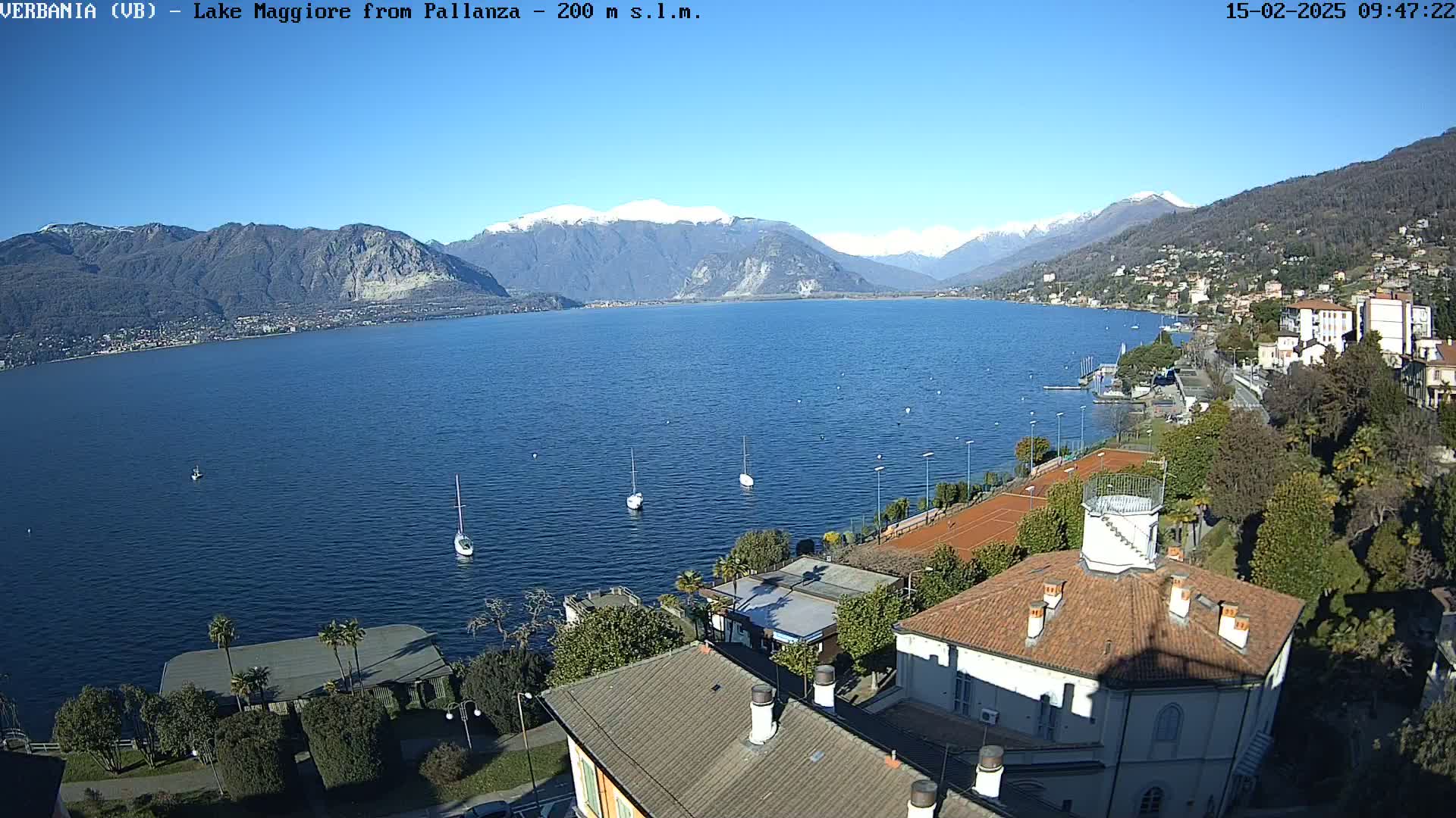 A clear, sunny day overlooking a lake with snow-capped mountains in the background, several sailboats on the water, and a town with buildings and tennis courts along the shoreline.