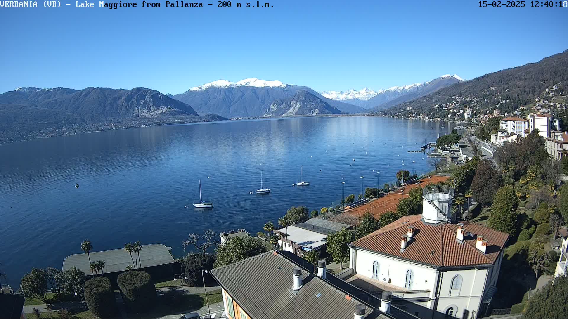 A clear, sunny day shows a view of a lake with snow-capped mountains in the background, sailboats on the water, and buildings along the shoreline.