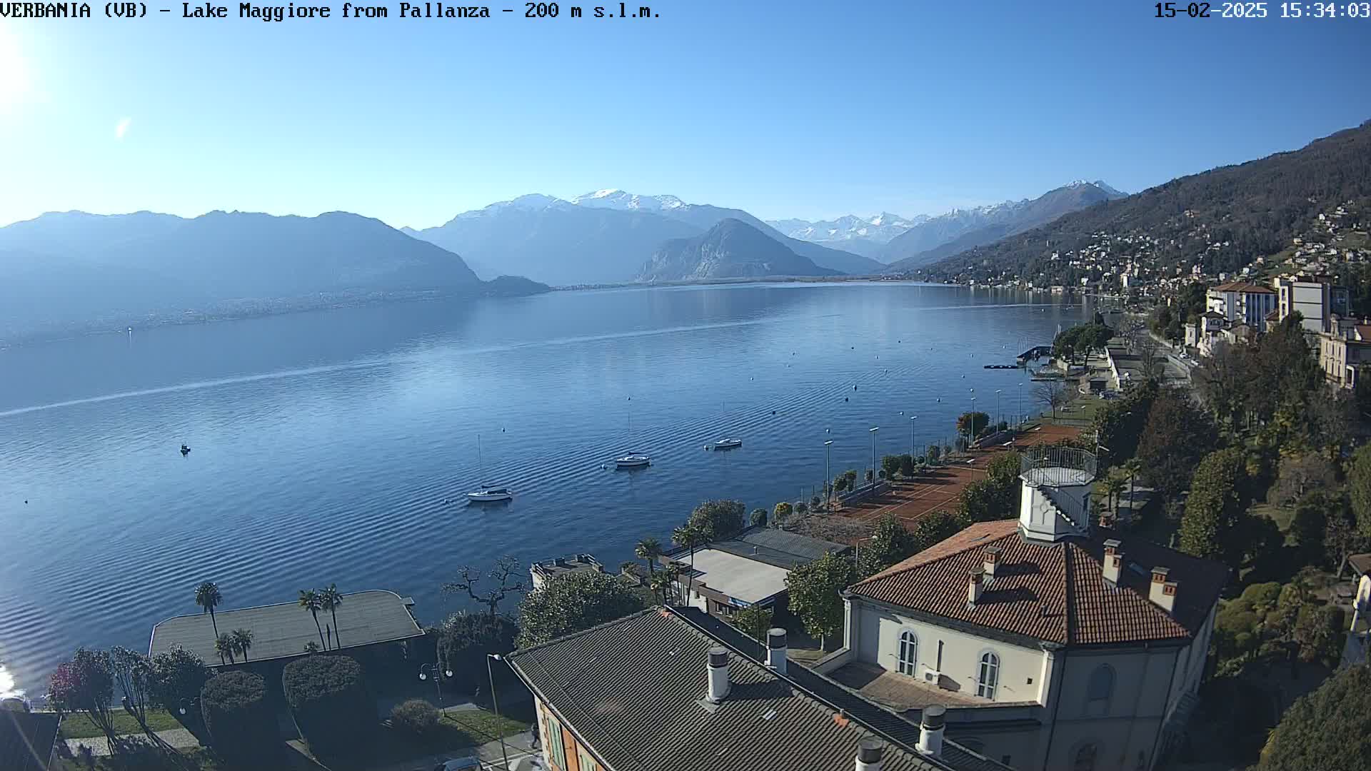 A calm lake stretches to snow-capped mountains under a clear blue sky, with several boats on the water and buildings along the lakeshore.
