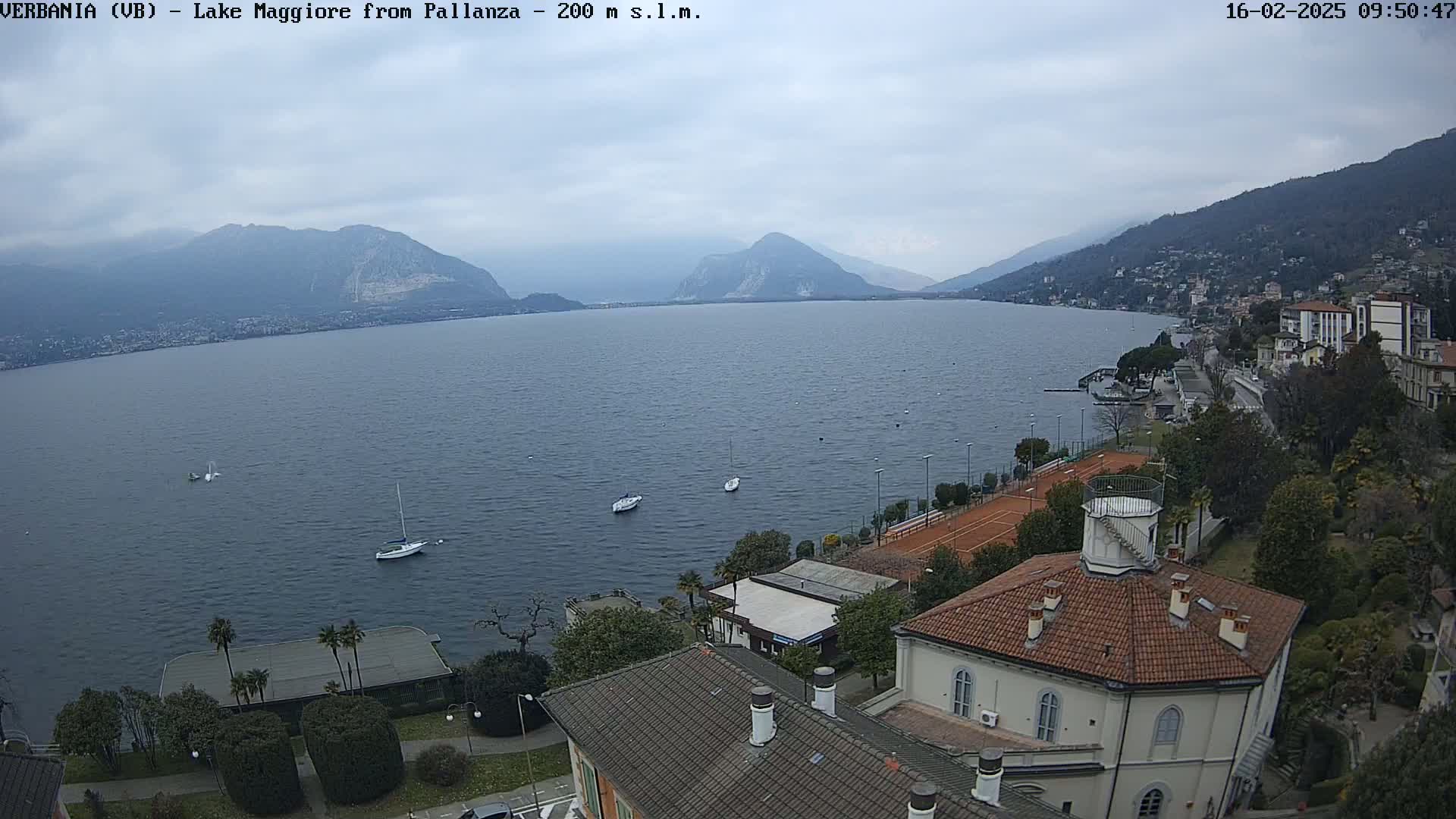 A view of Lake Maggiore from Pallanza on an overcast day, showing several boats on the water, buildings along the shoreline, and mountains in the background.