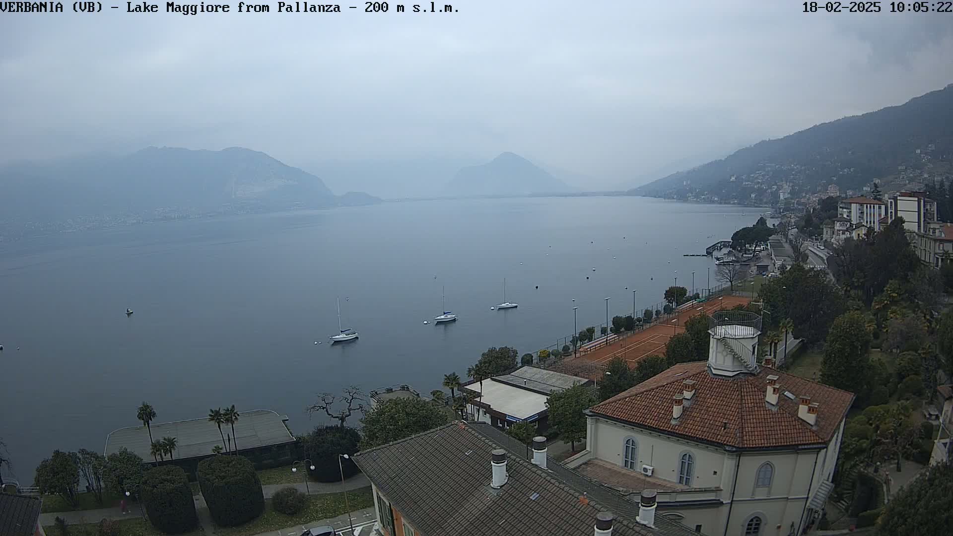 A hazy view of Lake Maggiore from a hillside, showing several sailboats on the water and a town with buildings and tennis courts below, under an overcast sky.