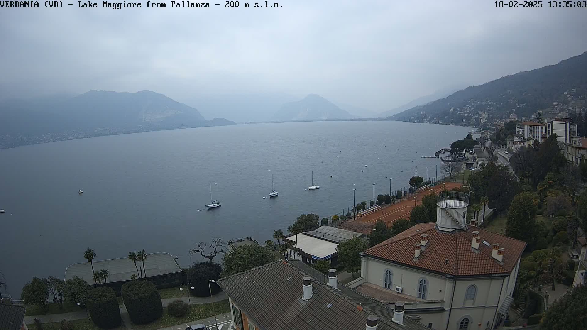 A hazy, overcast day reveals a tranquil lake scene with several sailboats, tennis courts, and buildings nestled along a shoreline, backed by mountains.