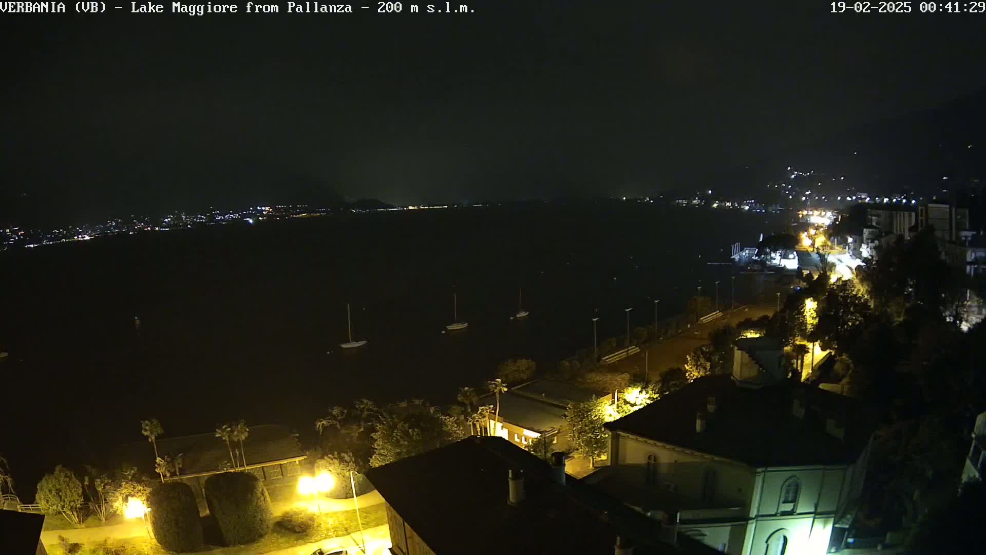 A nighttime view of a dark lake with several boats moored, a town with illuminated buildings lining the shore, and a calm, clear sky.