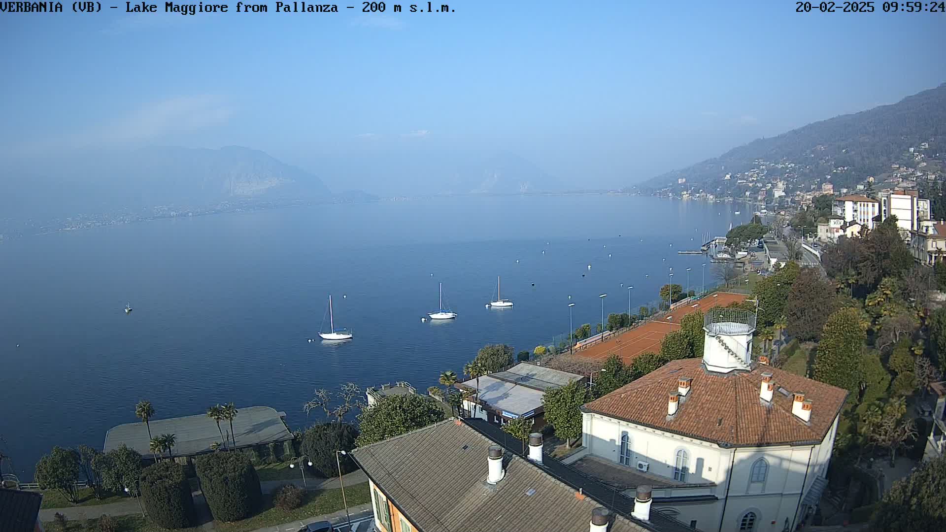 A clear, sunny day overlooks Lake Maggiore from a high vantage point, showing several sailboats on the water, a shoreline town, and tennis courts near a building with a red tile roof.