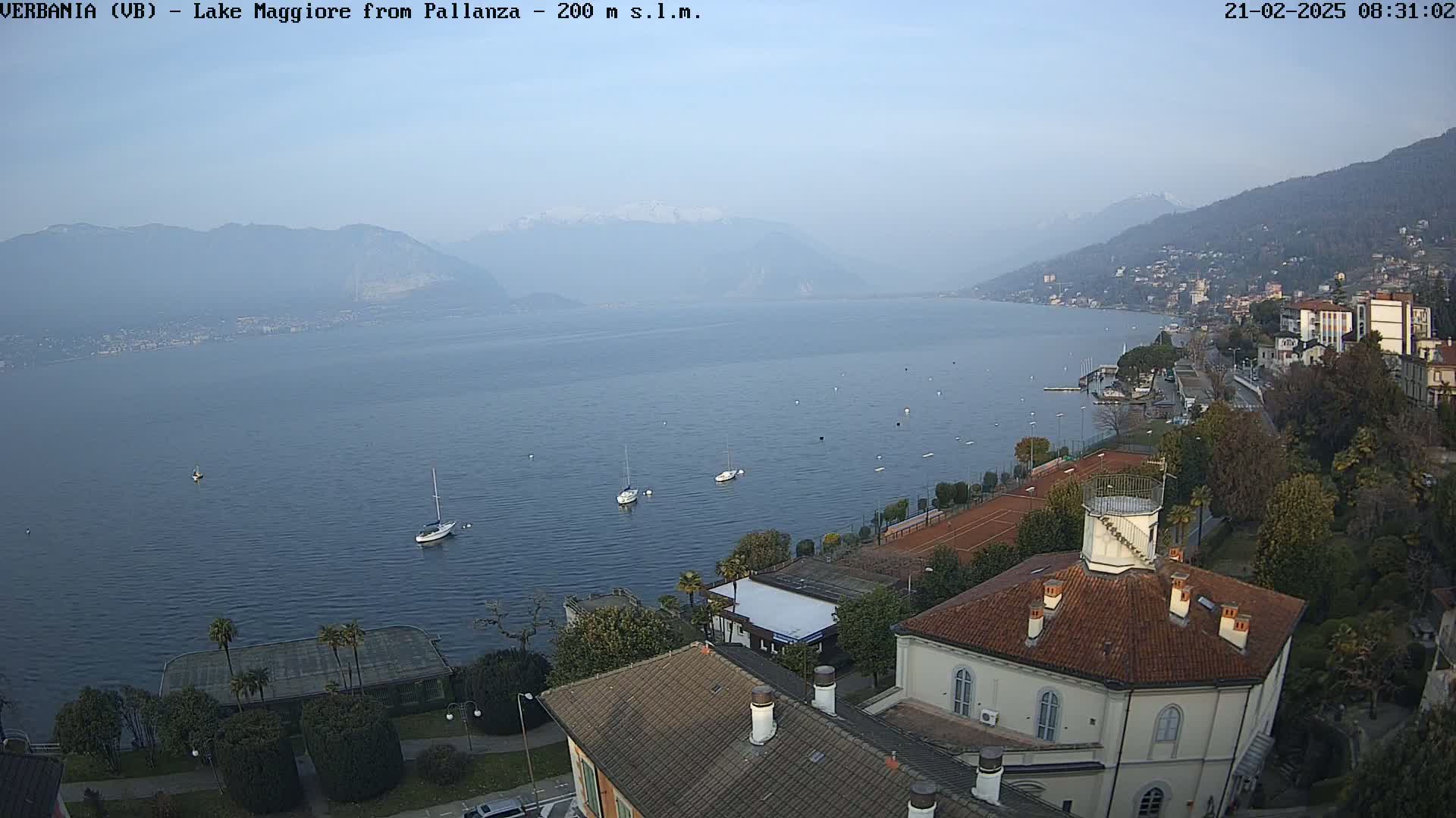 A hazy view of Lake Maggiore from a high vantage point shows several sailboats on the water, tennis courts, and buildings with red tile roofs nestled amongst greenery, all under a clear, light-blue sky.
