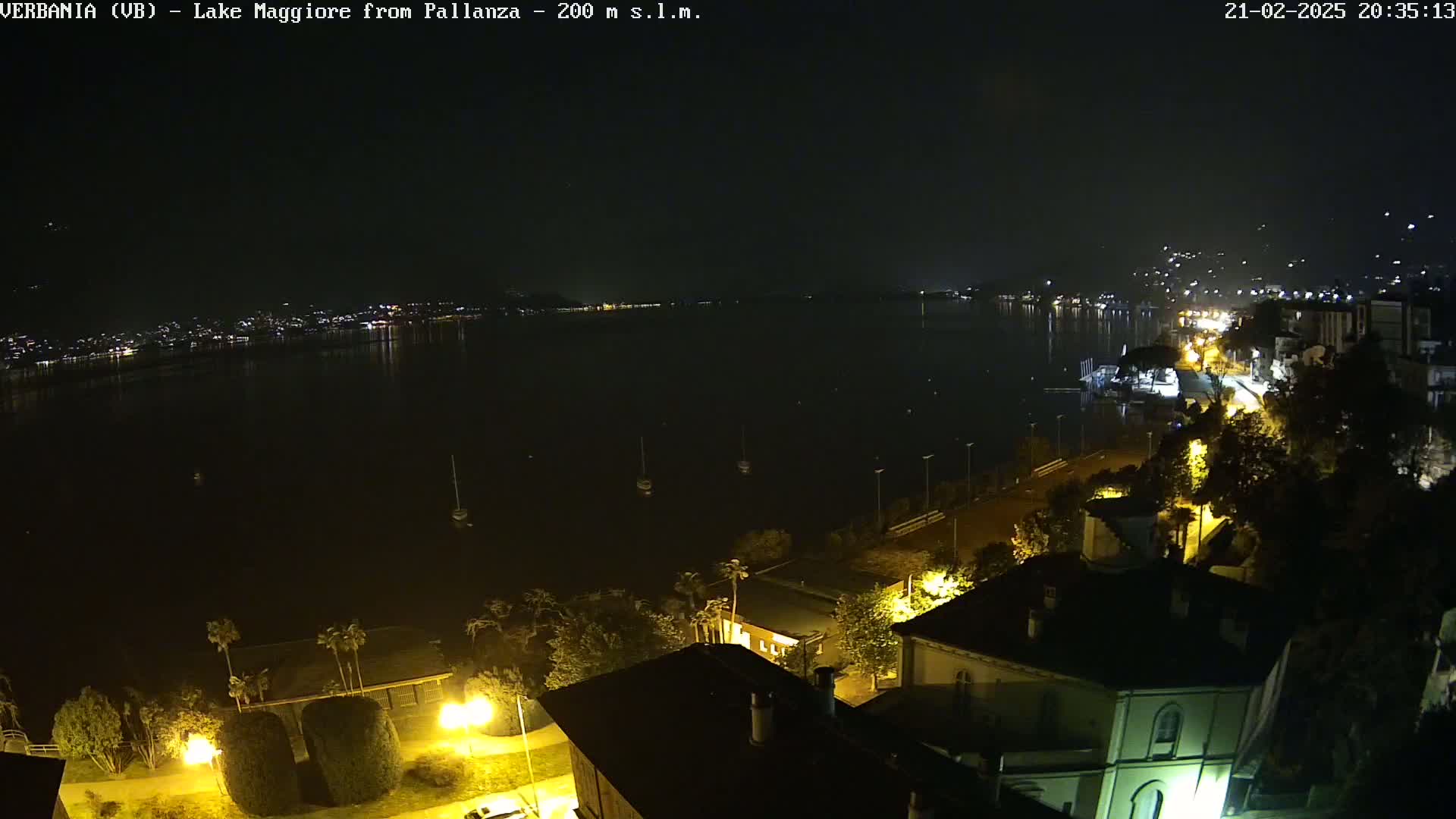 A nighttime view of a lake with a town's lights visible across the water and a few boats on the water, under a dark, clear sky.