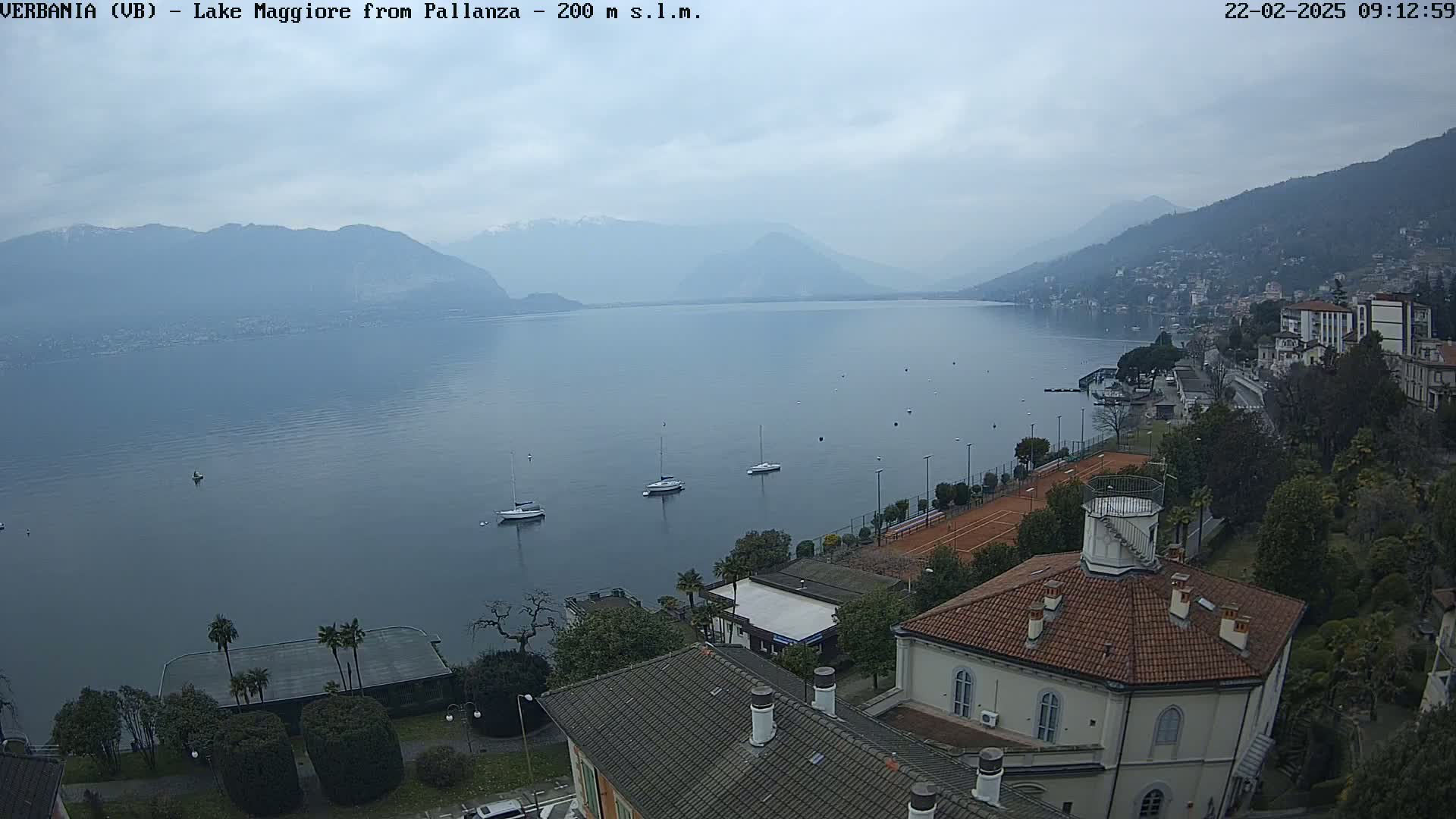 A calm lake with several sailboats is seen from an elevated vantage point on a cloudy day, overlooking a town with buildings and tennis courts nestled along the shoreline, backed by mountains.