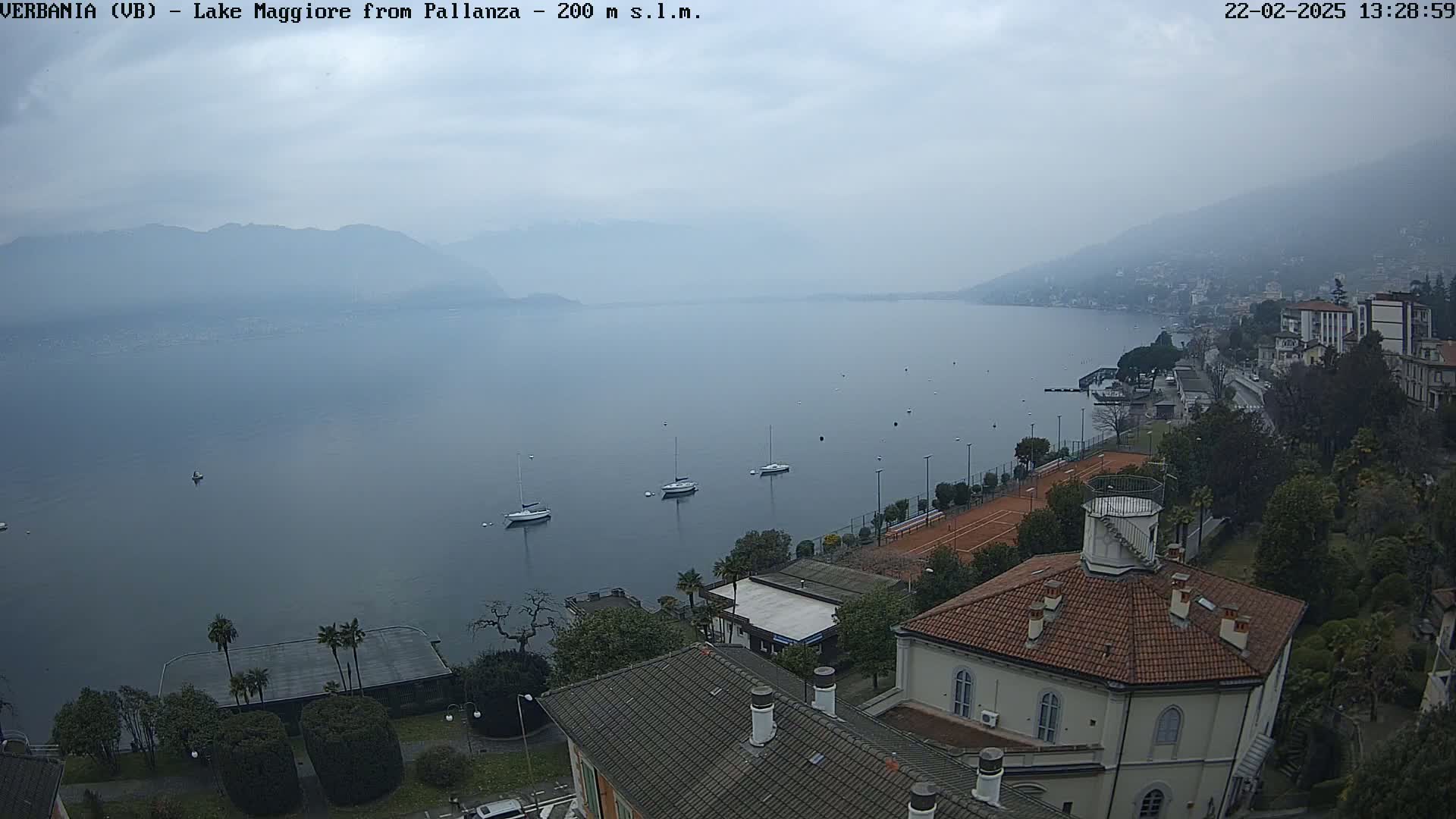 A hazy view of Lake Maggiore from a lakeside town shows several sailboats on the water, tennis courts, and buildings with red-tiled roofs under a cloudy sky.
