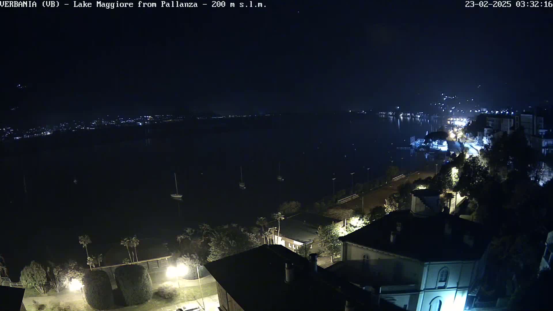 A nighttime, high-angle view of a dark lake with a few boats, and a shoreline with buildings and streetlights illuminated.