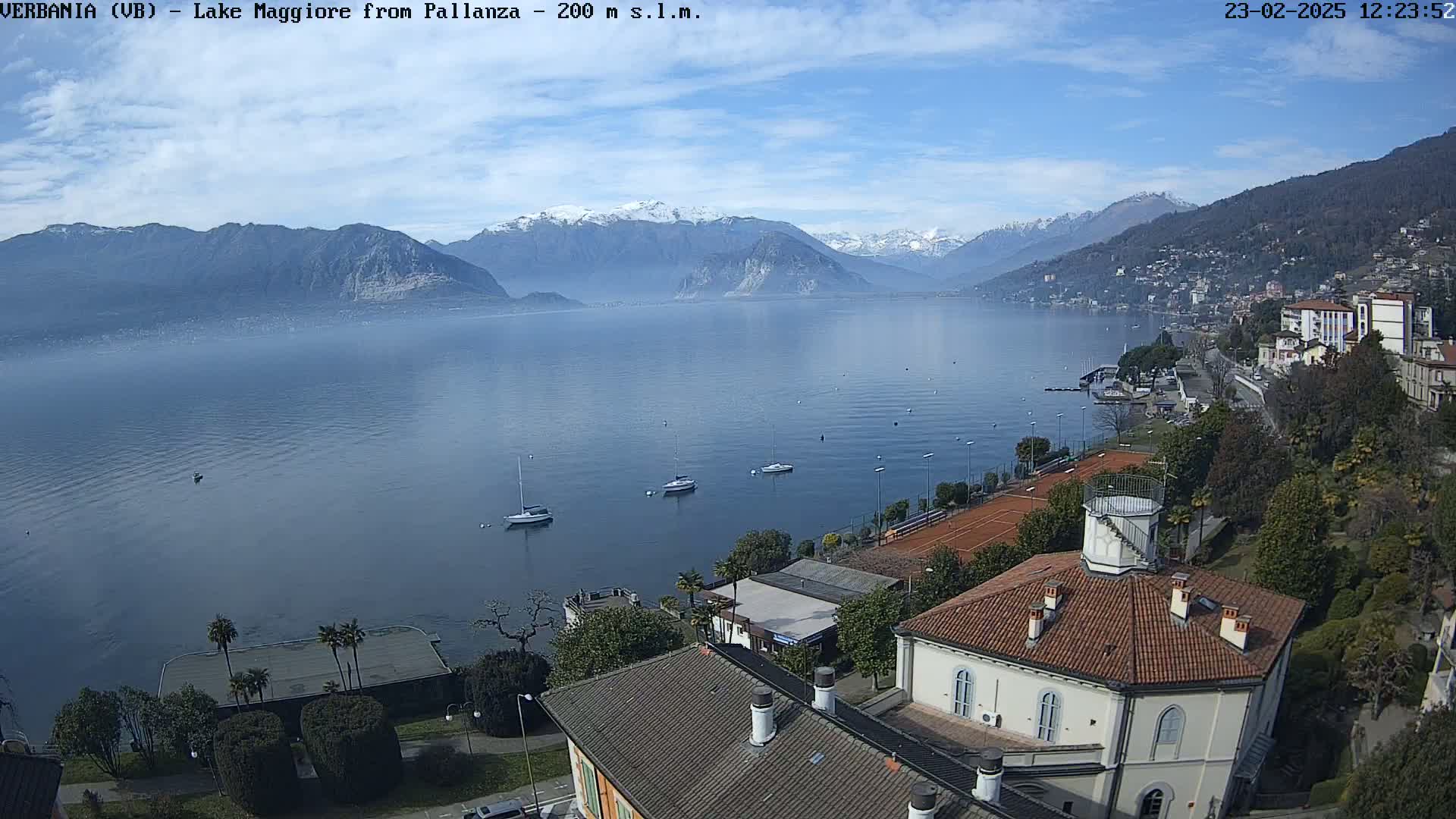 A clear, sunny day overlooking a lake with several sailboats, tennis courts, and buildings on the shore, with snow-capped mountains in the distance.