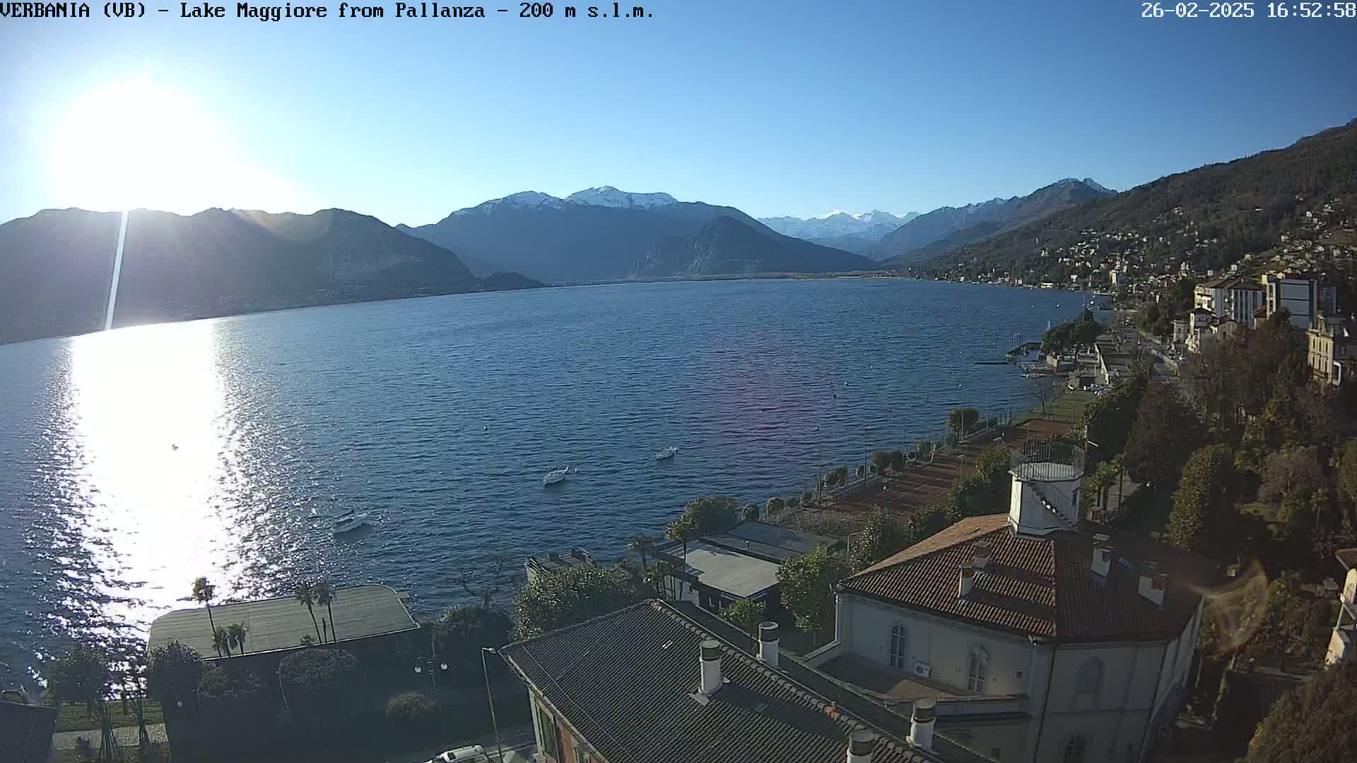 A sunny day shows a wide expanse of calm blue water reflecting sunlight, with snow-capped mountains in the distance and buildings along a shoreline.