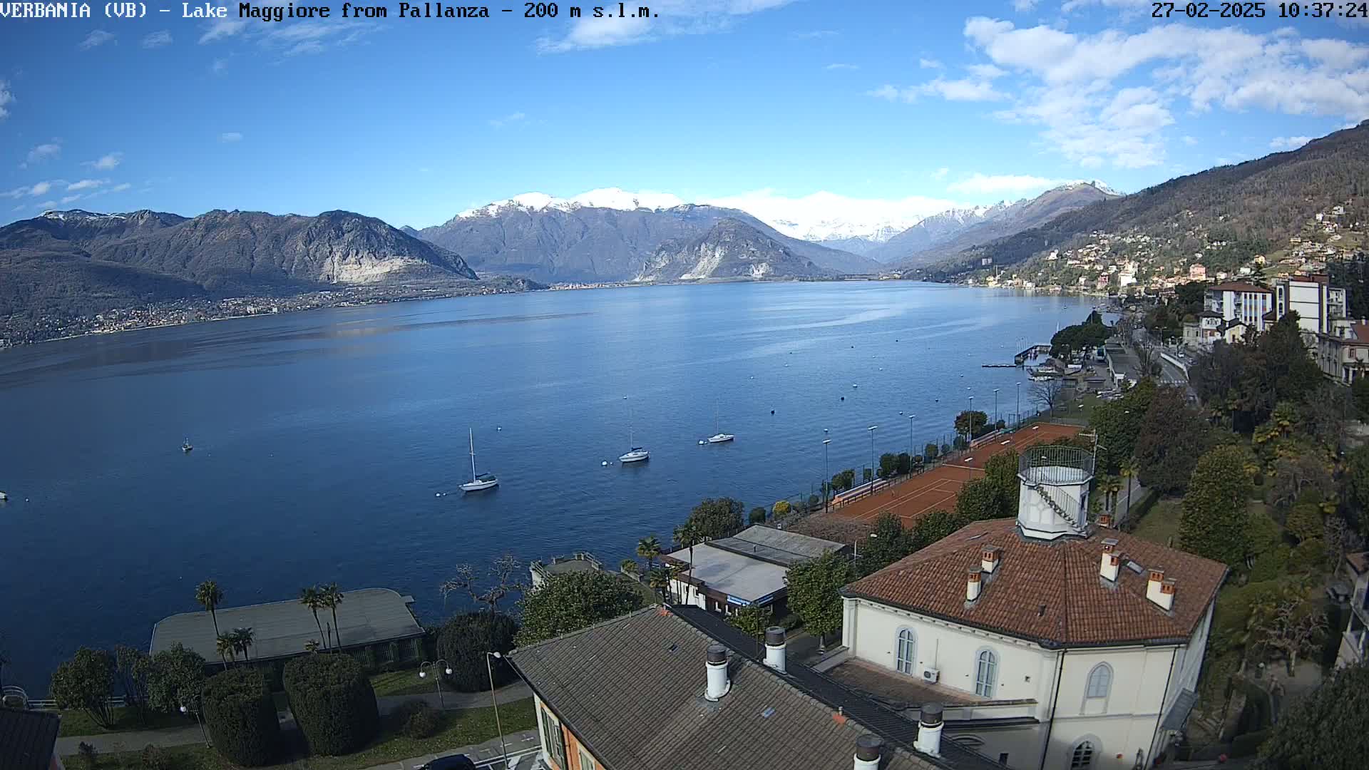 A lake with snow-capped mountains in the background, several sailboats on the water, and a town with buildings and tennis courts on the shore under a mostly sunny sky.