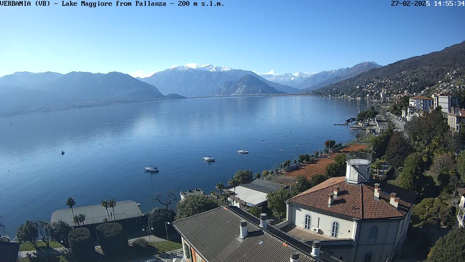A calm lake with several sailboats is seen from an elevated vantage point overlooking a town nestled against mountains, under a clear sunny sky.