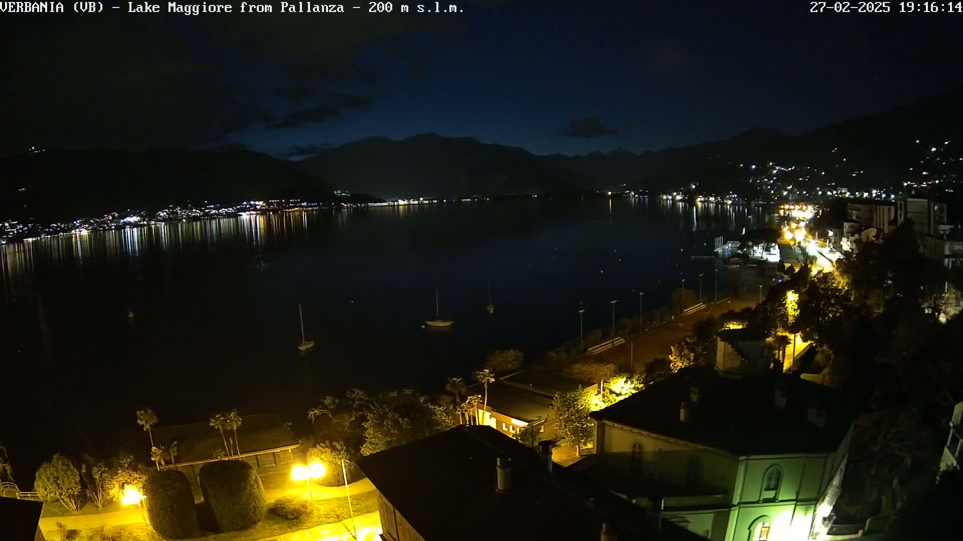 A nighttime view of a calm lake reflecting the lights of a town nestled against dark mountains under a clear, dark sky.