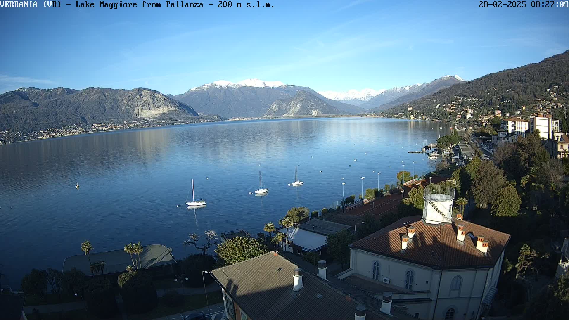 A clear, sunny day overlooks a lake with snow-capped mountains in the background; several sailboats are visible on the water near a shoreline town.