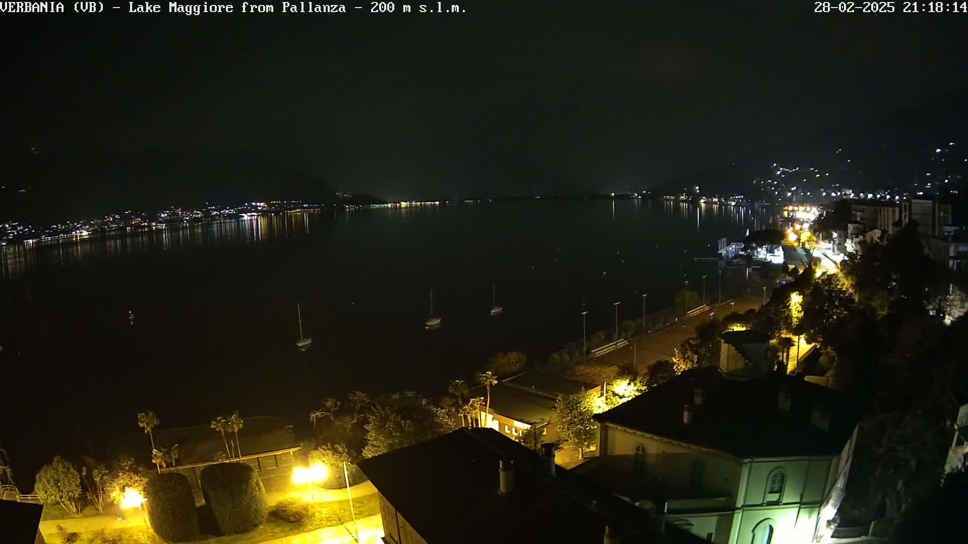 A nighttime view of a calm lake reflecting the lights of a city across the water, with a shoreline lined with buildings and trees illuminated by streetlights.