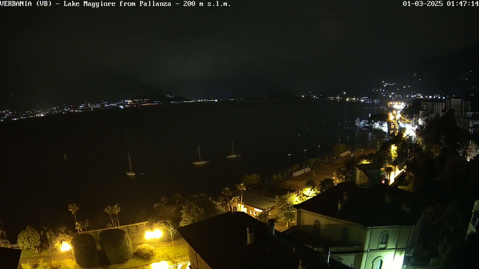 A nighttime view of a lake with a few boats and a town's lights visible across the water, under a dark, cloudy sky.