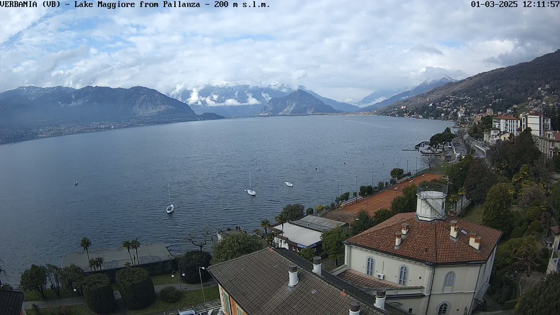 A view of Lake Maggiore from Pallanza, showing snow-capped mountains in the distance under a partly cloudy sky, with several sailboats on the lake and buildings along the shoreline.